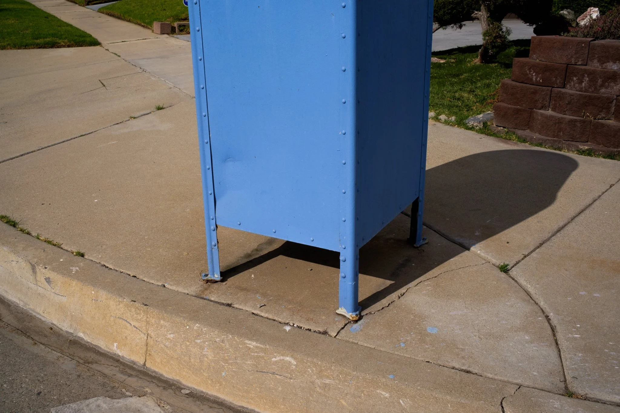 Blue utility box on a sidewalk with grass, brick wall, and sidewalk in the background, casting a shadow.