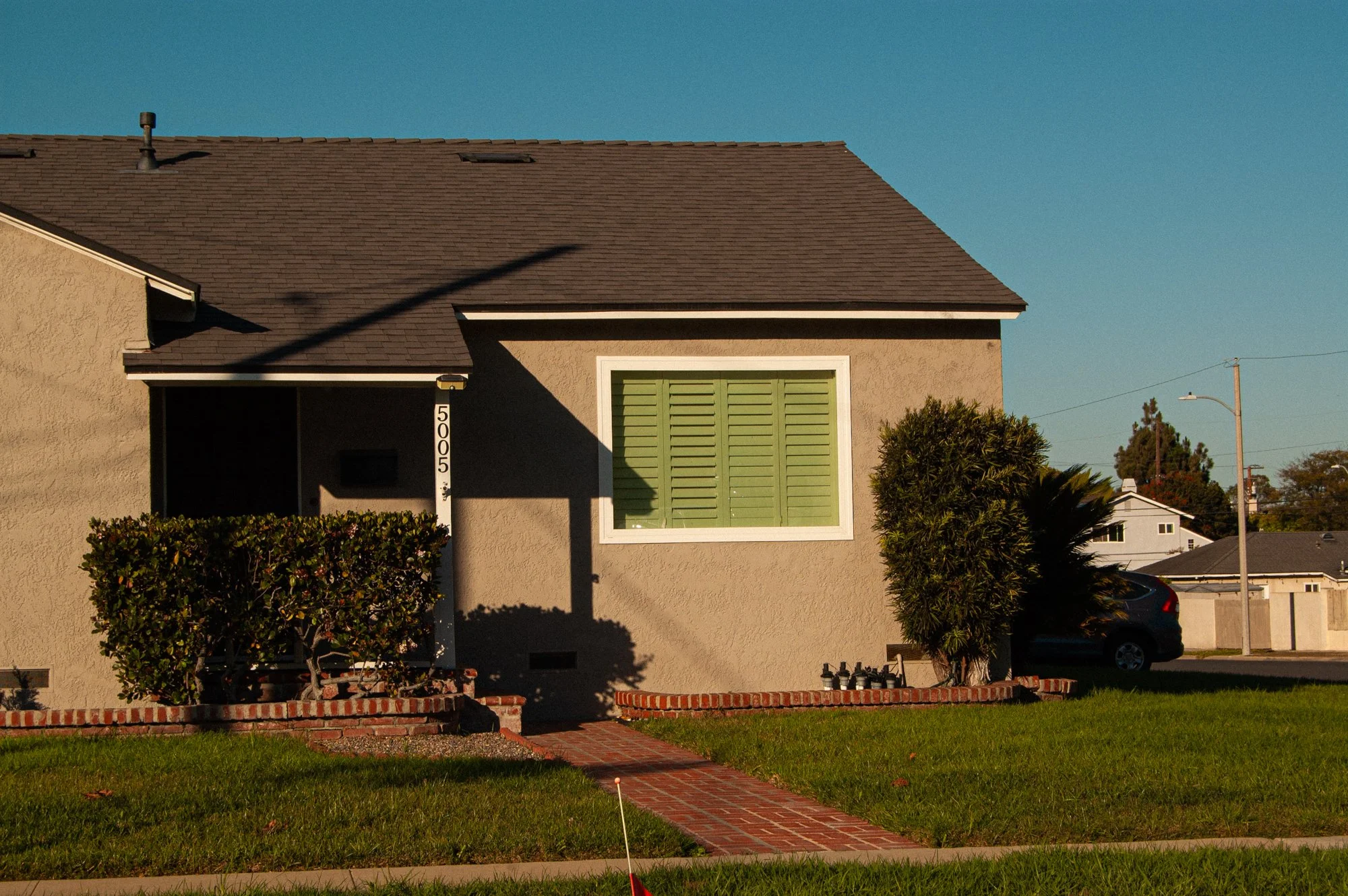 A two-story house with beige stucco walls, green shutters on a window, and a brown shingle roof. There are bushes and a brick pathway leading to the house, with a lawn in the front. A parking lot with cars and a streetlamp are visible in the backgrou