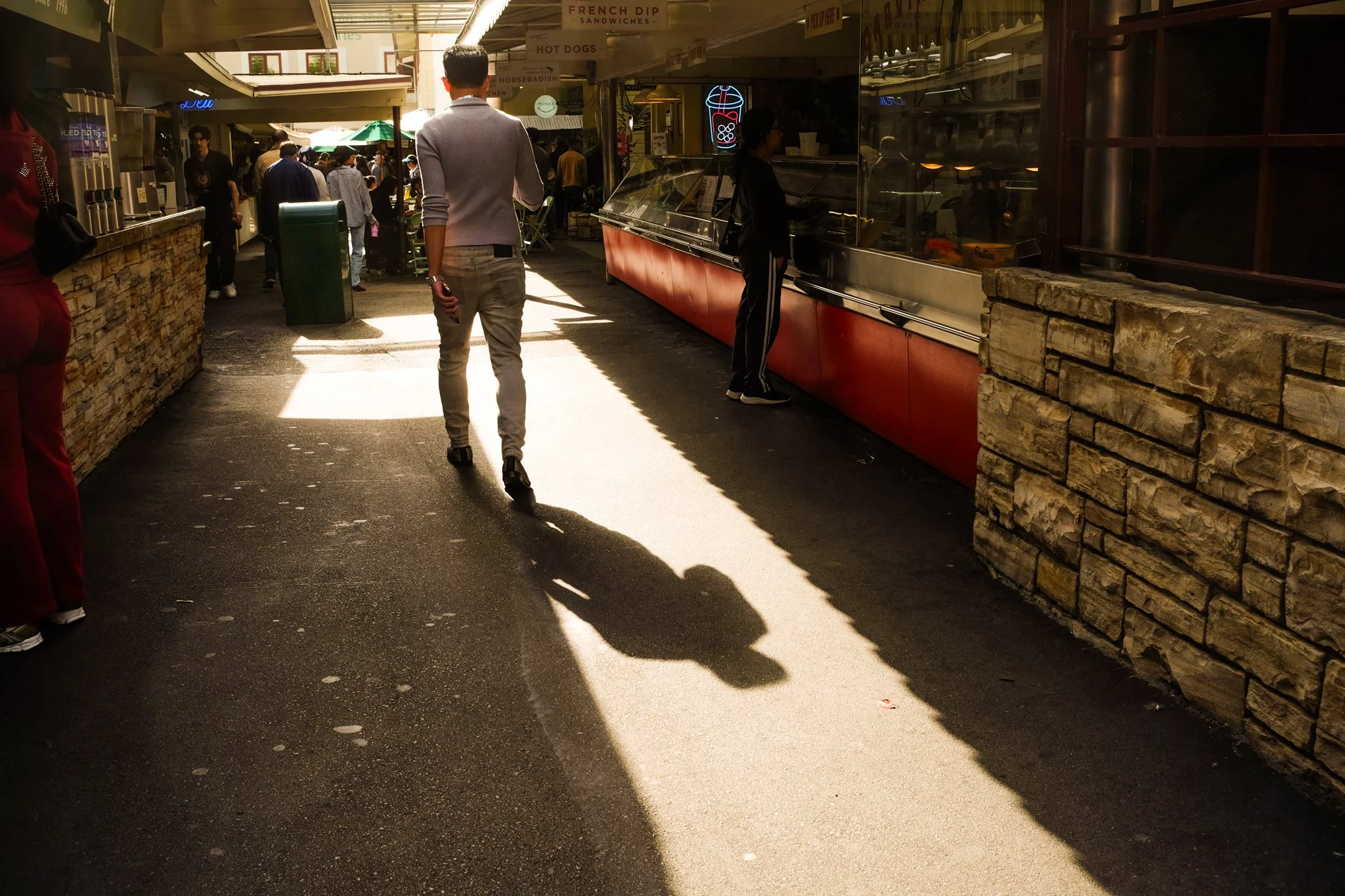A busy outdoor market or food court with various shops and vendors, including a counter with a red and black exterior, people walking and shopping, and signs for food options like French dip sandwiches and hot dogs.