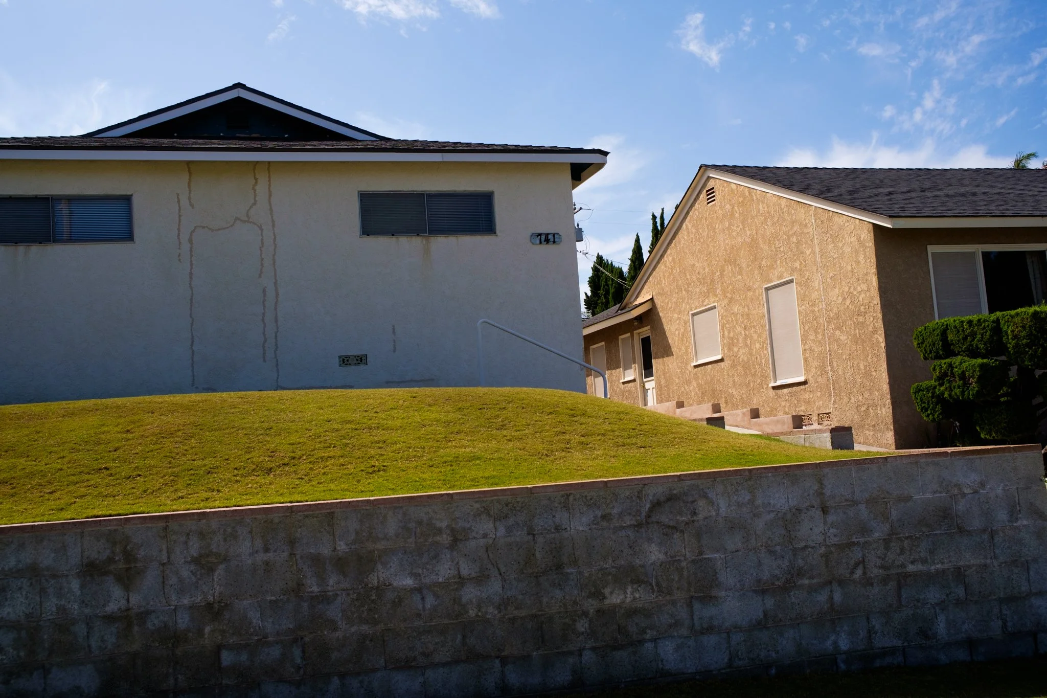 Two houses with stucco exteriors, one yellow and one beige, with sloped roofs against a cloudy blue sky. The yellow house has visible water stains on its wall. The foreground features a small grassy hill and a stone retaining wall.