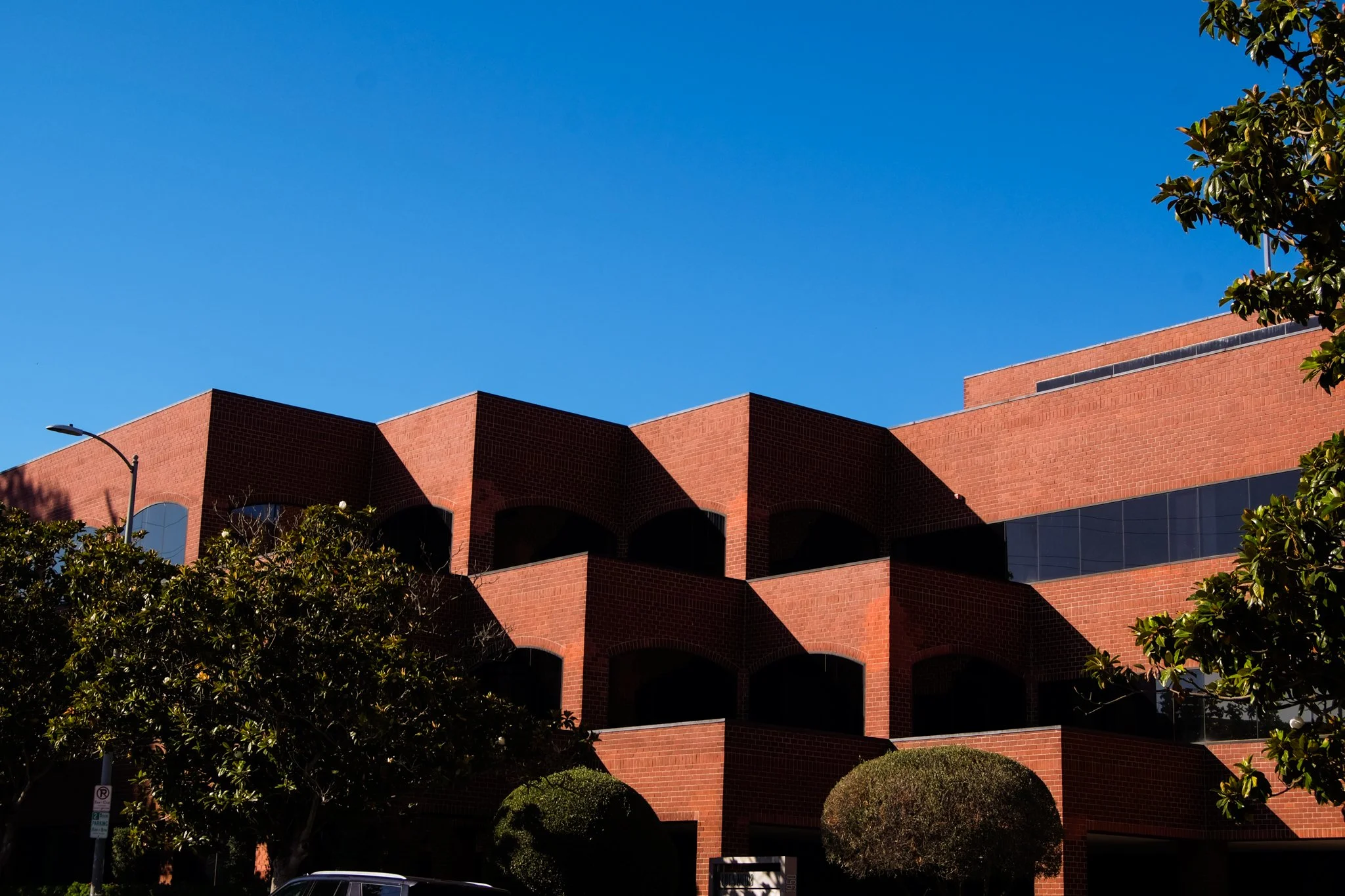 A modern brick building with arched windows, partially obscured by trees and bushes, under a clear blue sky.
