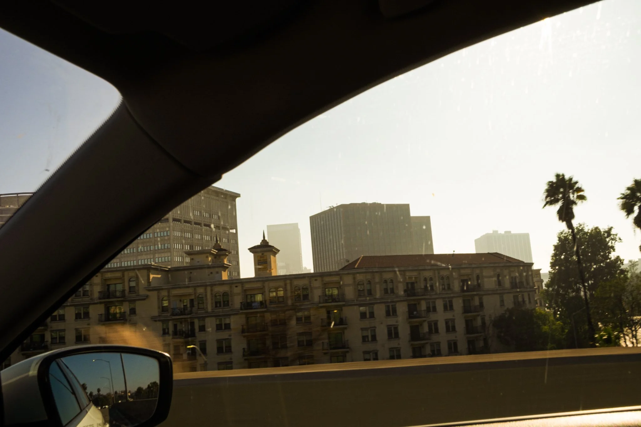 Cityscape viewed from a car window, including tall buildings, palm trees, and an ornate architectural building in the foreground, with a rearview mirror reflecting part of the scene.