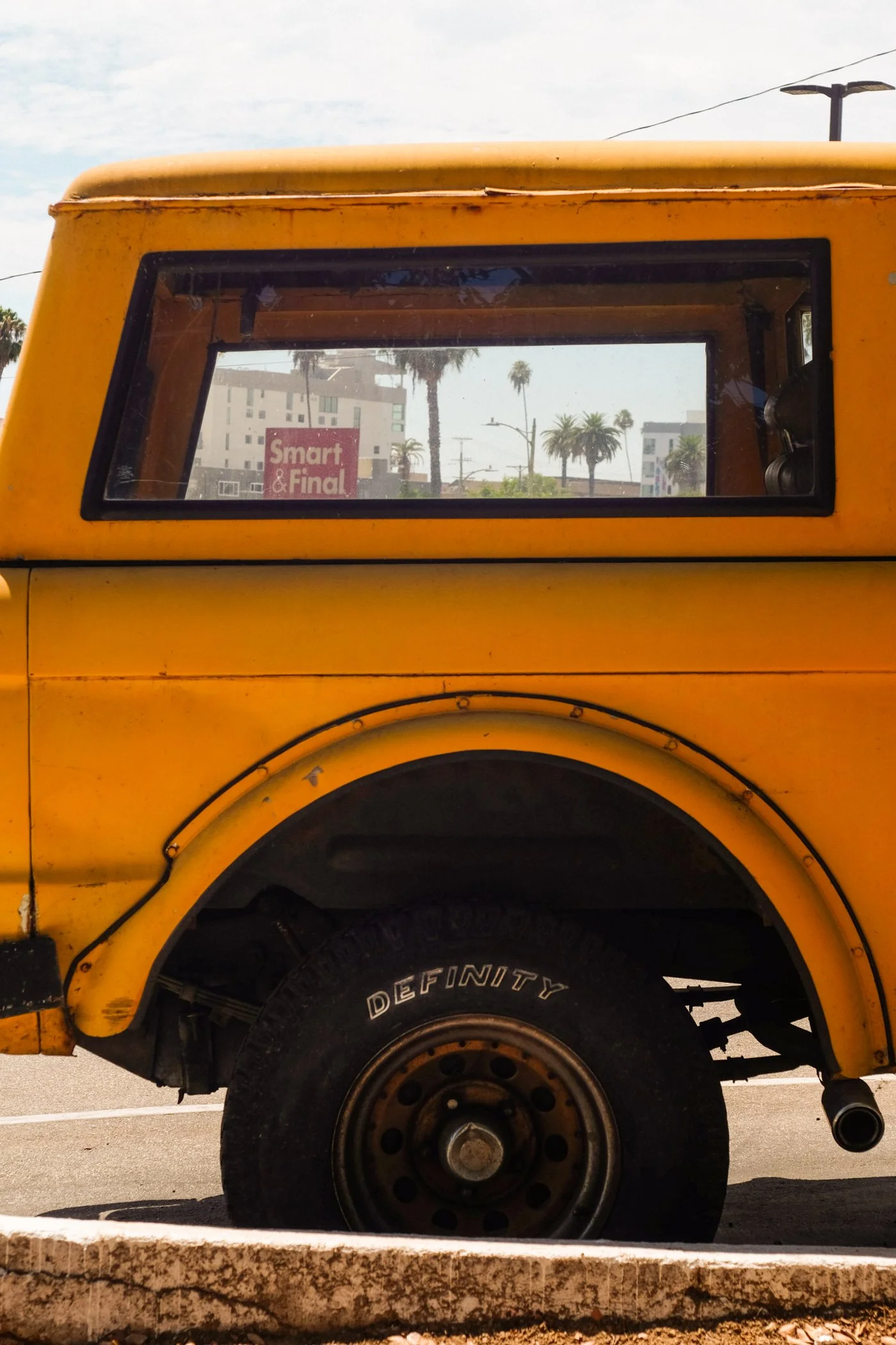 Close-up of a yellow vehicle, focusing on the side and wheel. The window area shows palm trees and buildings in the background, with a sign that reads 'Smart & Final' visible through the window.