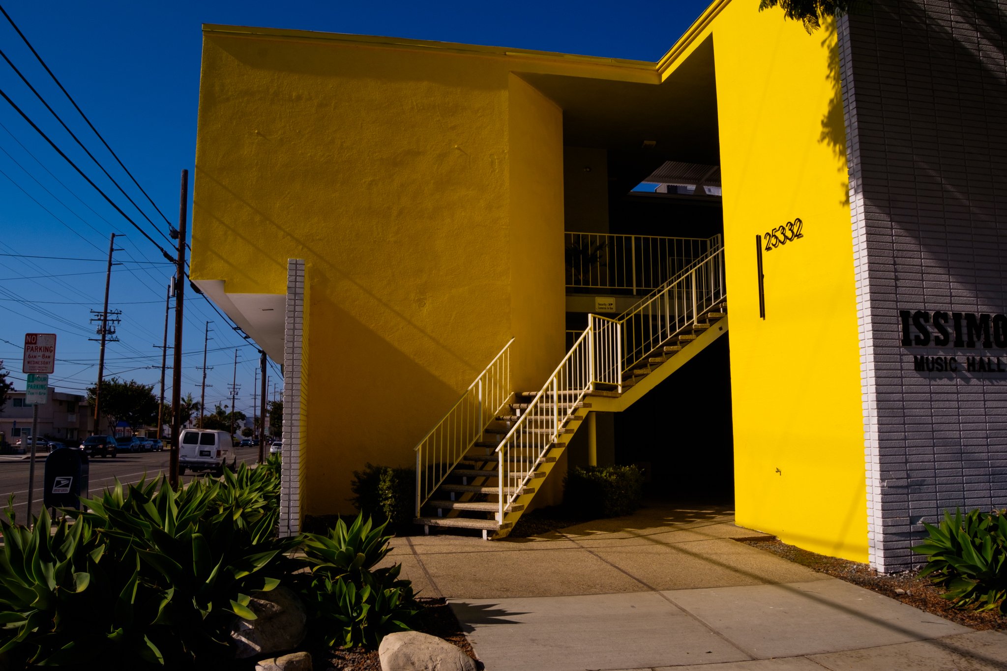 A yellow building with a staircase on the outside leading to an upper floor, with the number 25332 on the wall, and part of a sign that reads 'ISSIMO.' The building is near a street with power lines and parked cars, under a blue sky.