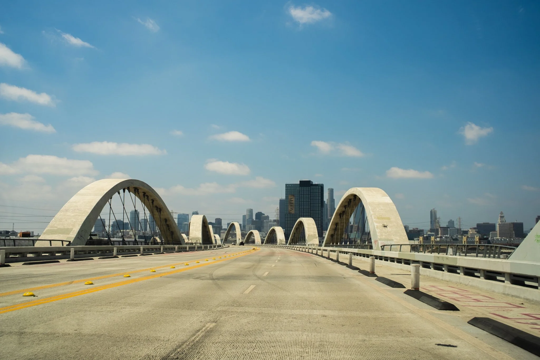 View of a bridge with multiple concrete arches over a highway, with a city skyline in the background under a partly cloudy blue sky.
