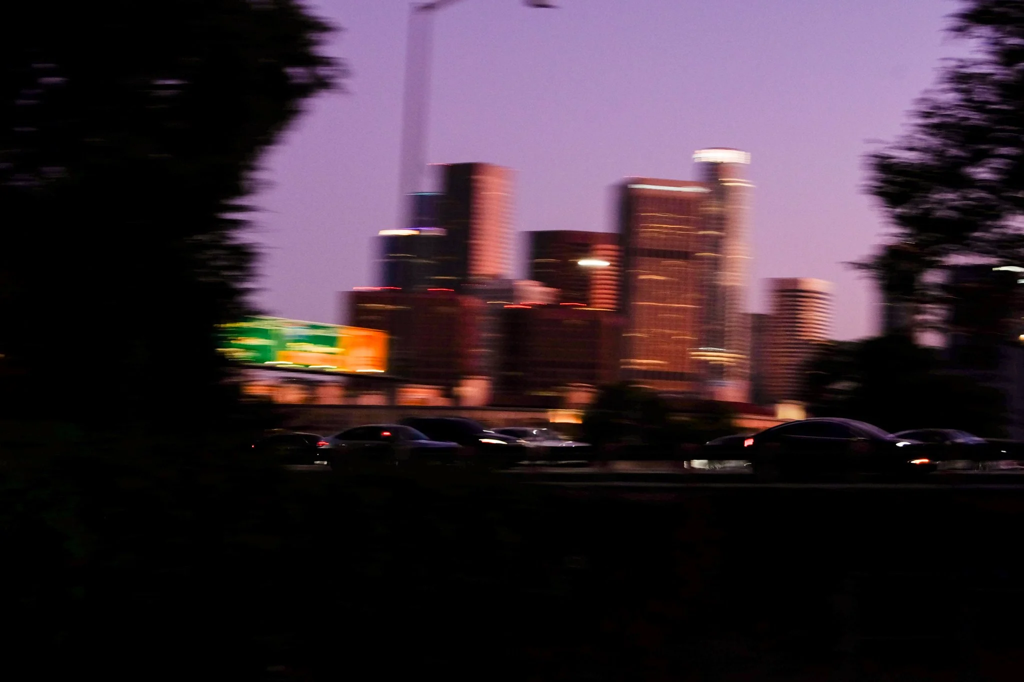 A blurry cityscape at dusk with tall buildings, some with illuminated tops, and a purple sky. There are trees on both sides of the image and a row of dark cars parked at the bottom.