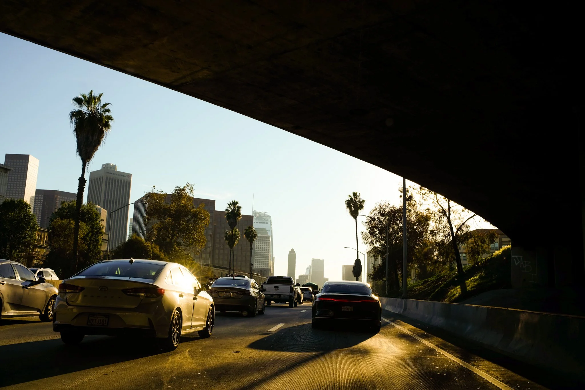 City street viewed from underneath an overpass, with parked cars in the foreground, tall buildings, trees, and clear sky in the background, during sunset.