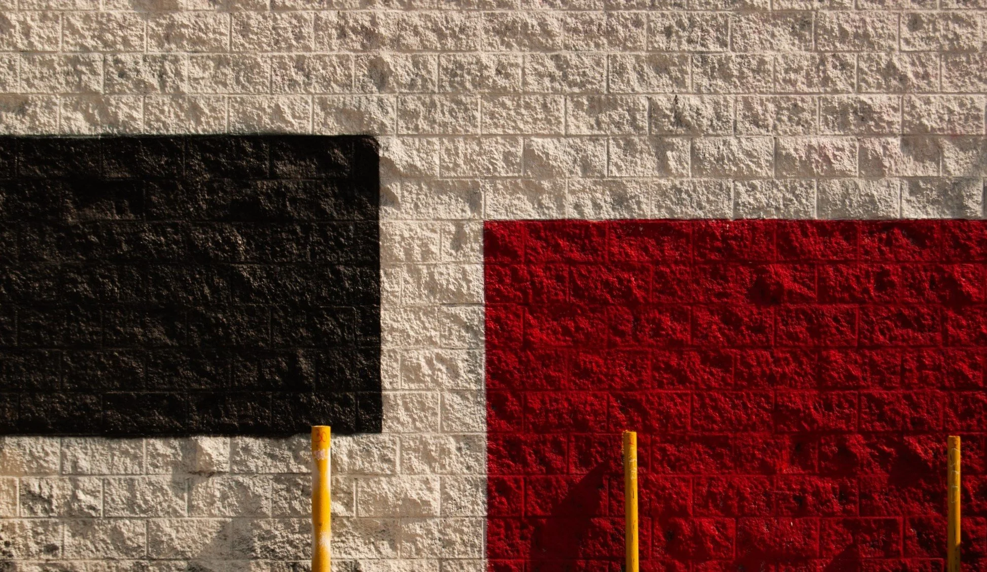 A brick wall painted with the Texas flag, featuring black, white, and red vertical stripes. Three yellow posts are in front of the wall.