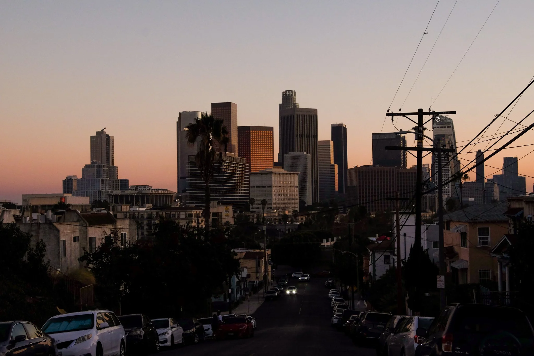 Sunset city skyline with tall buildings and palm trees, viewed from a residential street with parked cars.