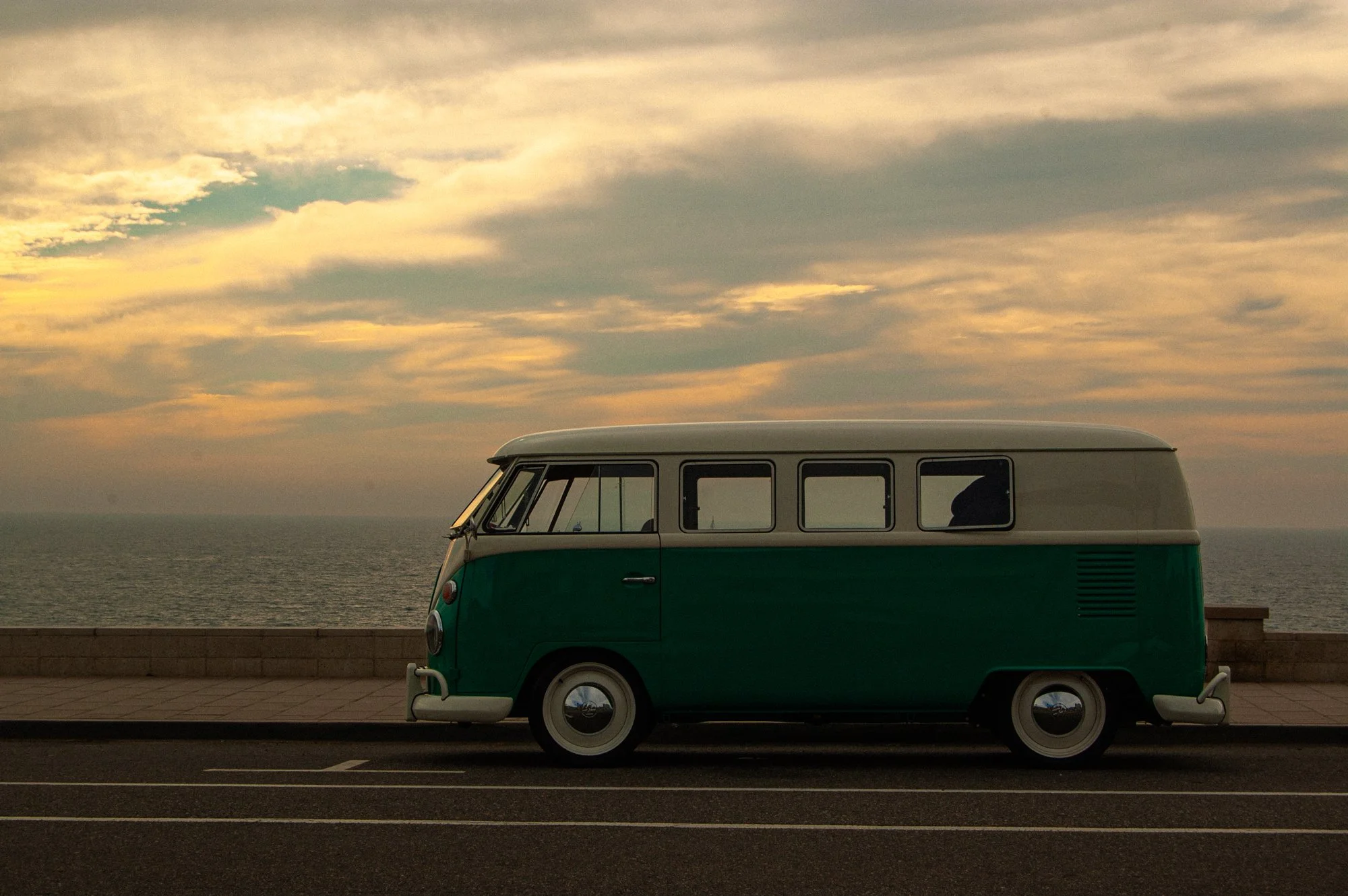 A vintage green and white Volkswagen camper van parked along a seaside promenade at sunset, with cloudy sky and calm ocean in the background.