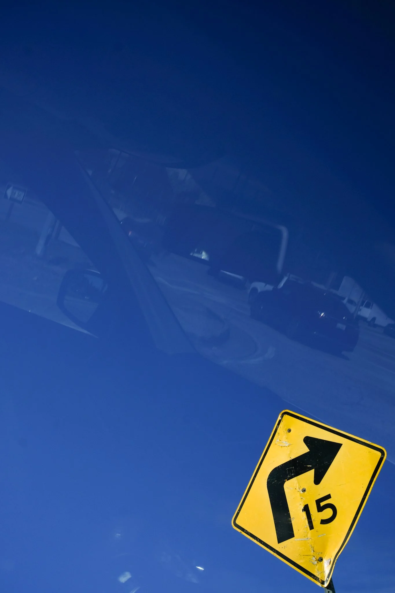 A yellow warning road sign indicating a sharp turn with a 15 mph speed limit. The sign is tilted, and there is a reflection of a vehicle and blue sky with some clouds on a shiny surface, possibly a car's windshield.