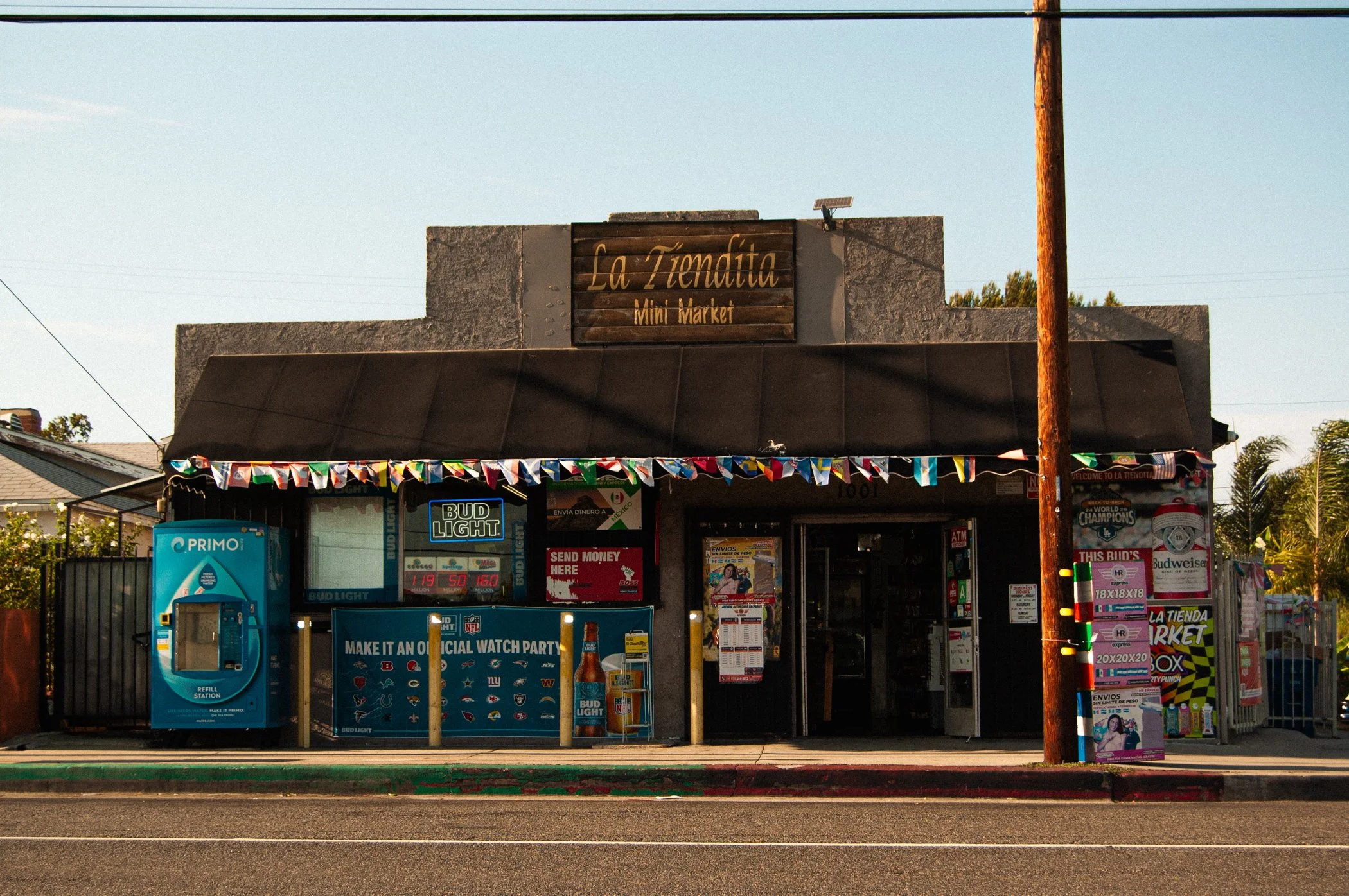 A small convenience store with a wooden sign that reads 'La Tiendita Mini Market.' The store has various advertisements and products visible outside, including a Bud Light sign, a Pride flag banner, and lotto or gaming posters. A utility pole is in t