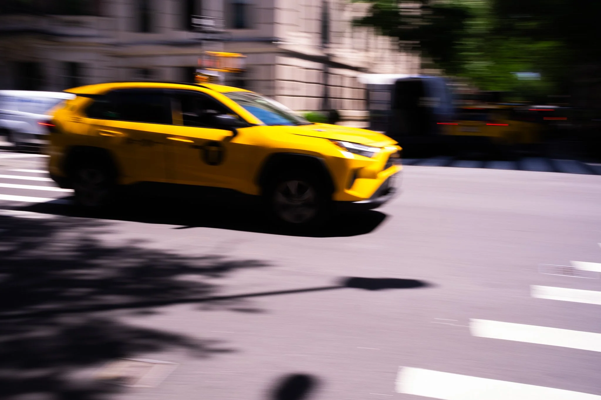 A yellow taxi cab moving quickly down a city street with blurred background buildings and trees.