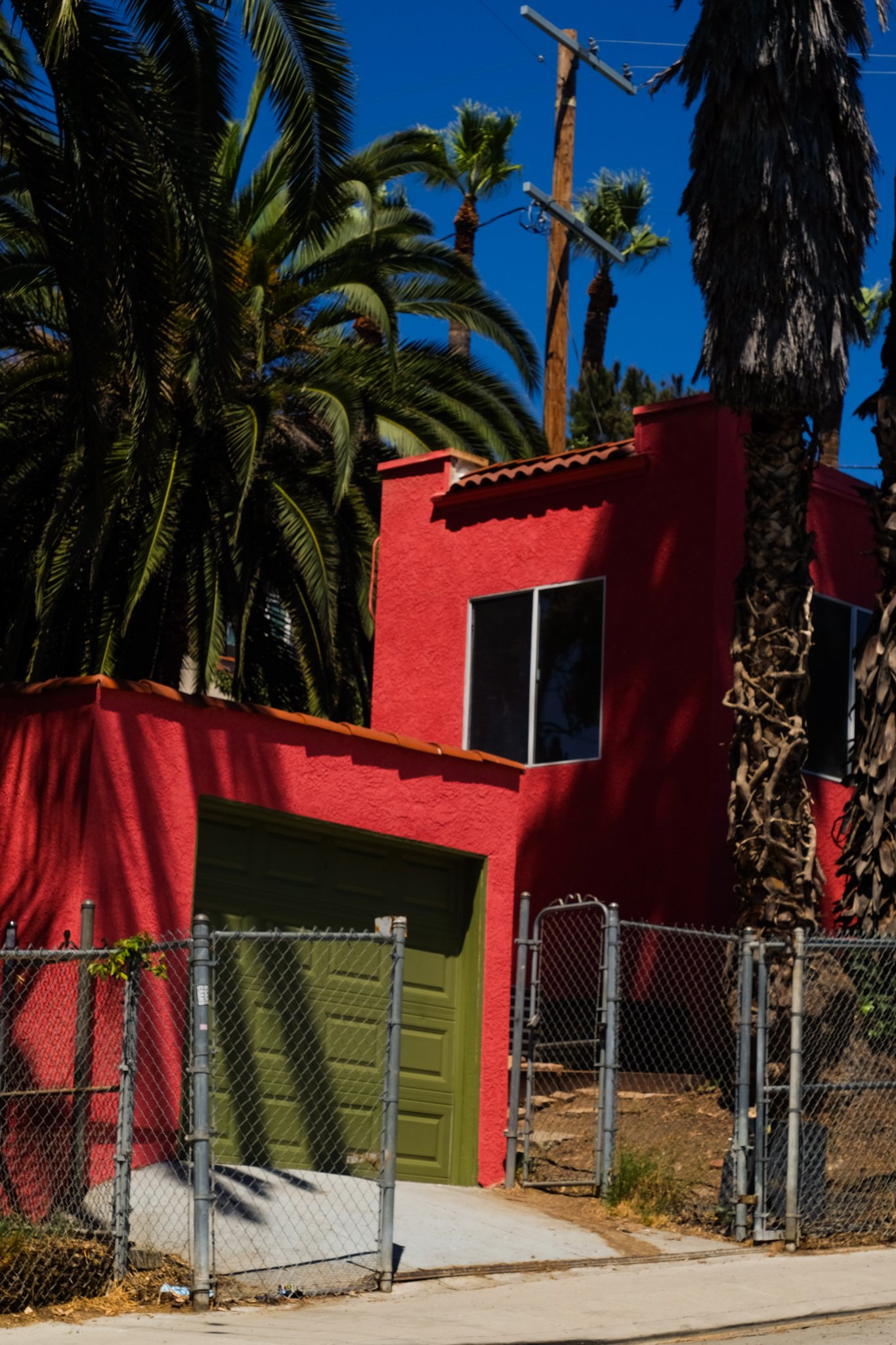 A red building with a green garage door, chain-link fence, tall palm trees, utility poles, and a blue sky.