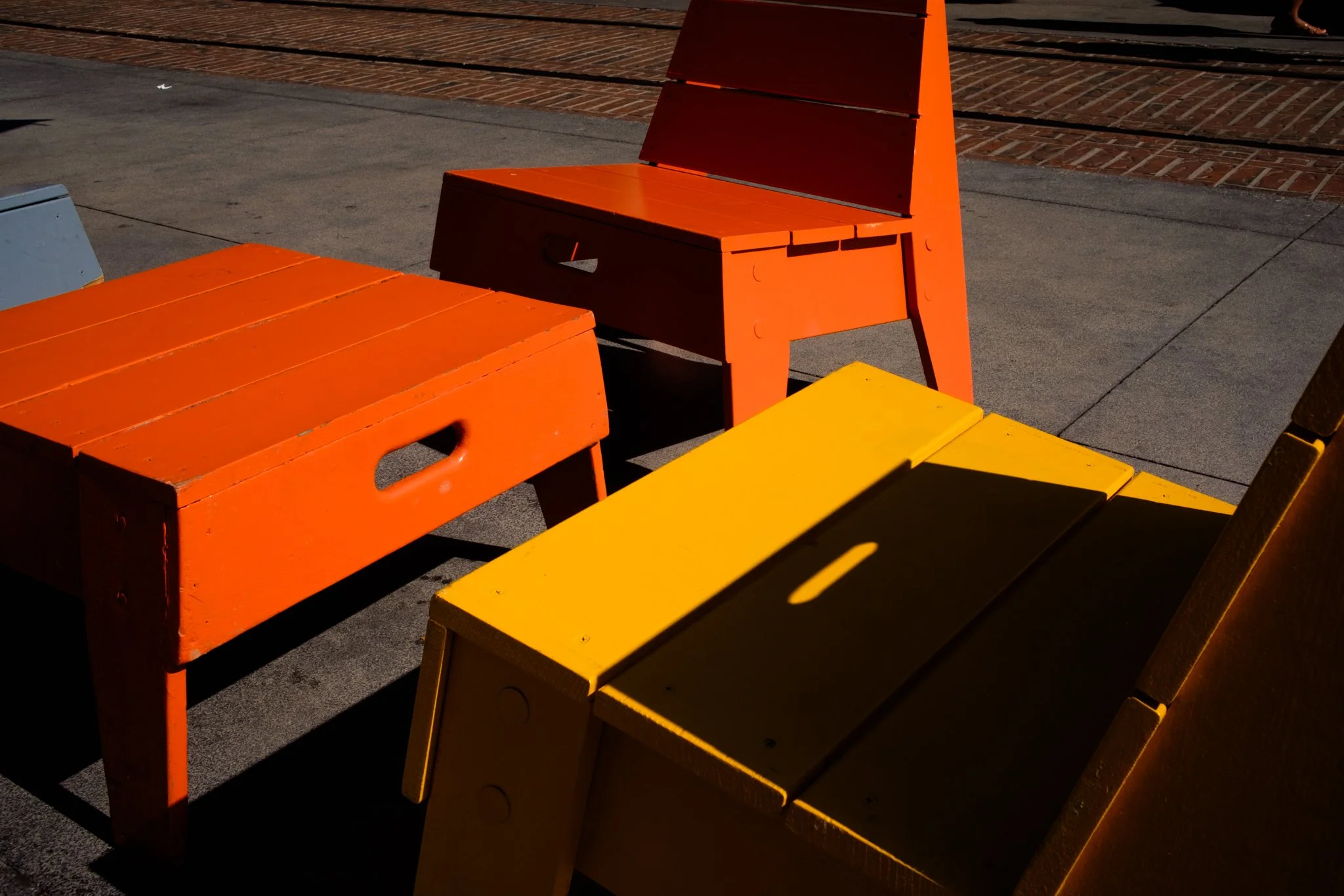 Multiple colorful benches, painted orange and yellow, placed on a concrete sidewalk with a brick surface in the background.