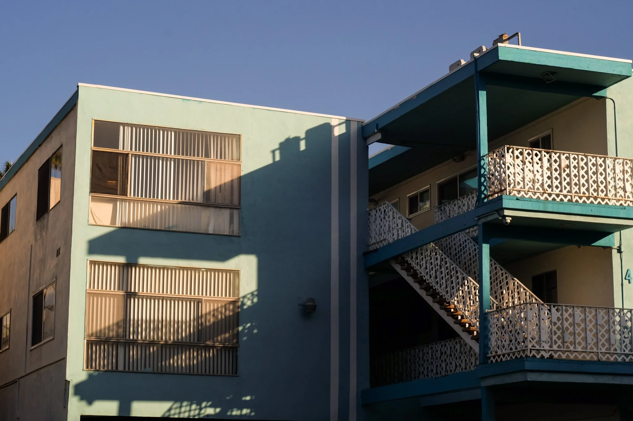 An apartment building with exterior stairs and balconies painted in pastel blue and green, with sunlight casting shadows on the facade.