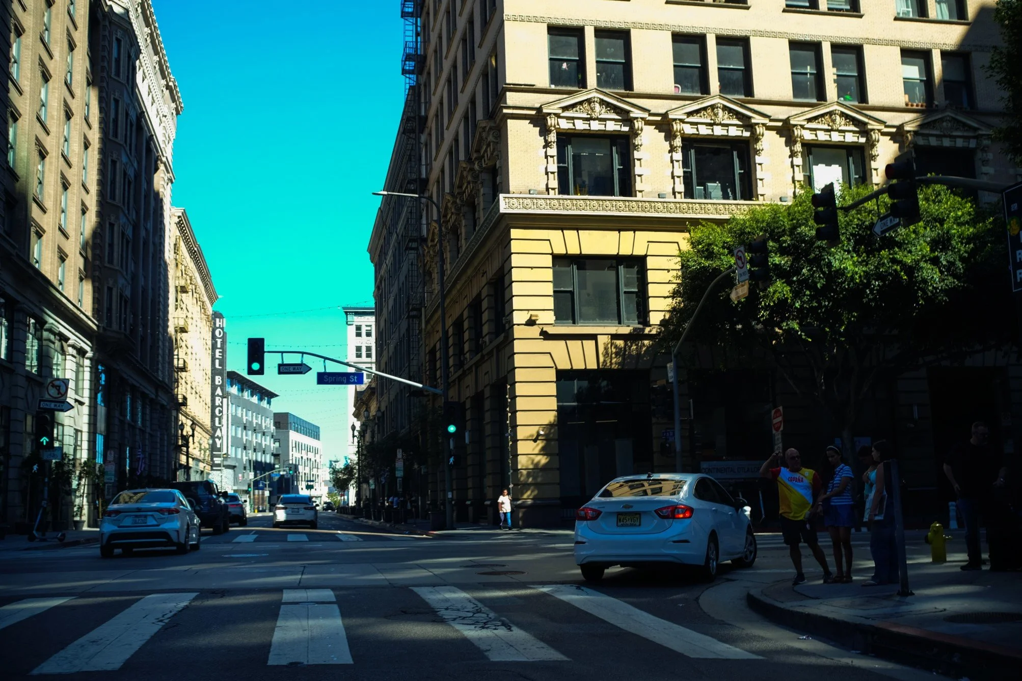 A city street intersection with tall historic buildings on both sides, pedestrians waiting to cross, cars stopped at a red light, and a bright blue sky overhead.