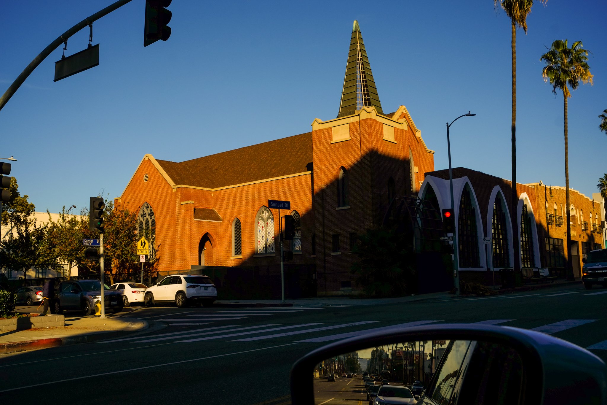 A brick church with a tall steeple and stained glass windows, surrounded by cars and palm trees on a sunny day.