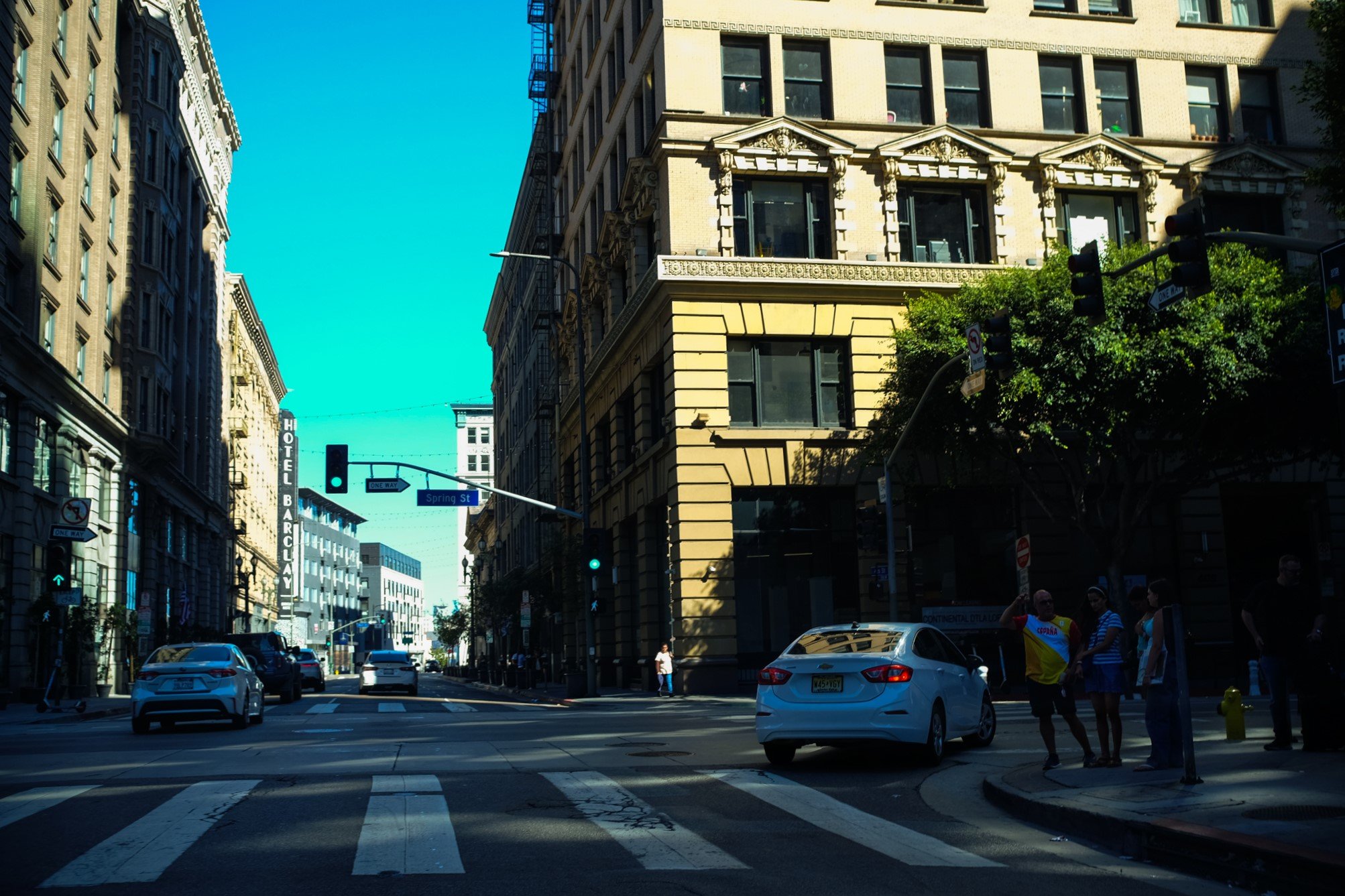City street with tall historical buildings on both sides, green traffic light, few cars on the road, a few pedestrians crossing, clear blue sky.