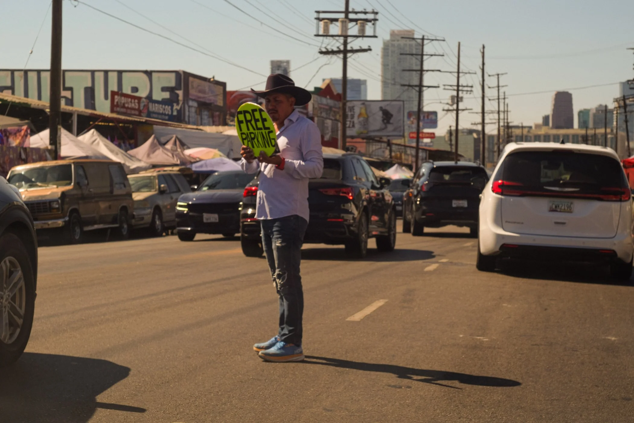 A man wearing a cowboy hat and light-colored shirt holding a bright green sign that says 'FREE PARKING' across a busy street with cars and urban storefronts in the background.