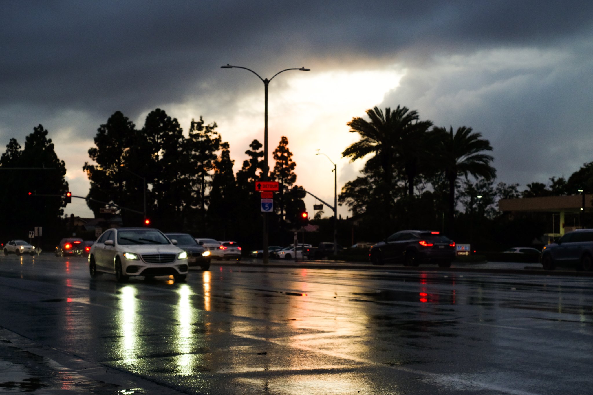 A city street scene during a storm or rain, with dark clouds in the sky and wet pavement reflecting the streetlights and cars. Several cars are visible driving on the road, and traffic lights are illuminated. Silhouettes of trees and street signs are