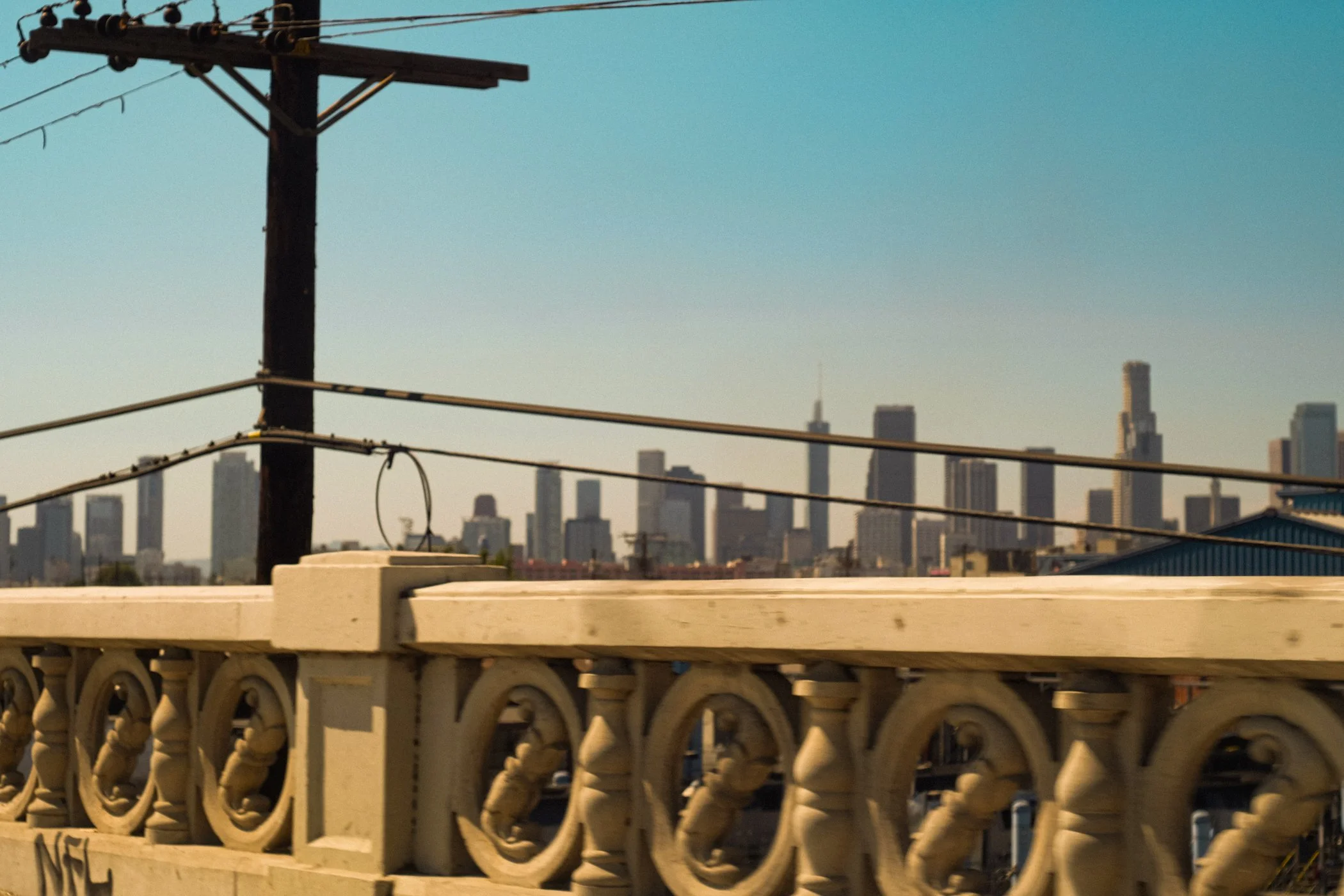 View of a city skyline with tall buildings in the background and a utility pole with wires in the foreground, seen from a bridge or elevated roadway.