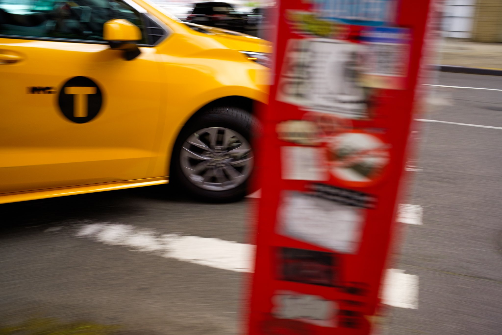 A yellow taxi cab passing by a red newspaper vending machine on an urban street.