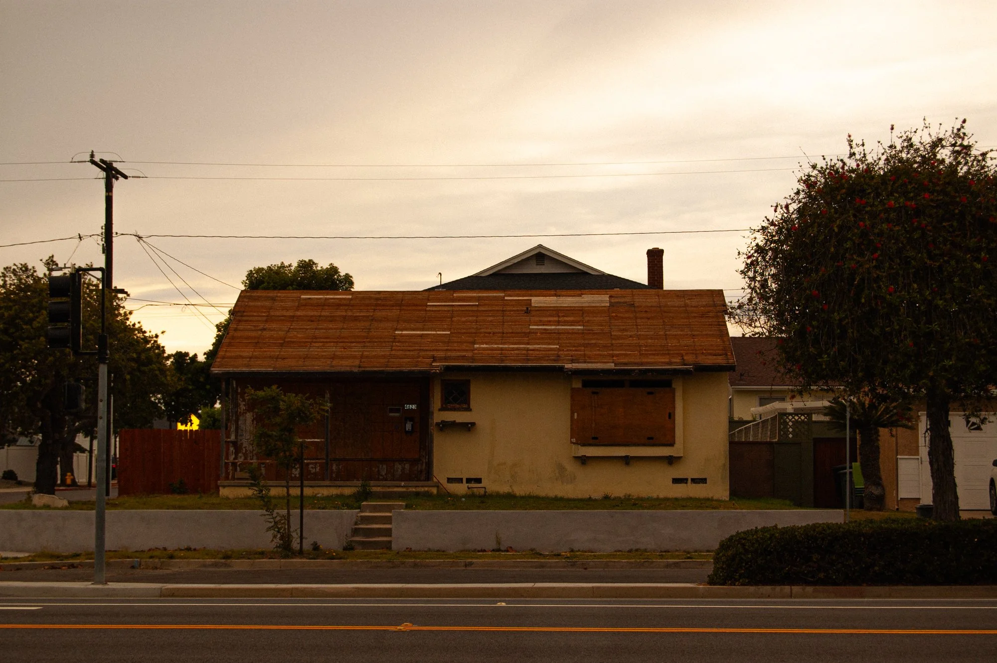 A residential street scene at dusk with a house that has boarded-up windows and a missing roof section, trees, a utility pole with power lines, a traffic light, and a sidewalk in the foreground.