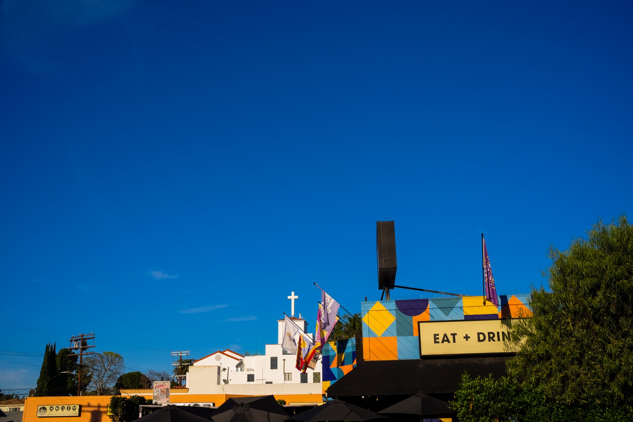 Colorful building with flags and a yellow sign that reads 'Eat + Drink', a cross in the background, and a bright blue sky.