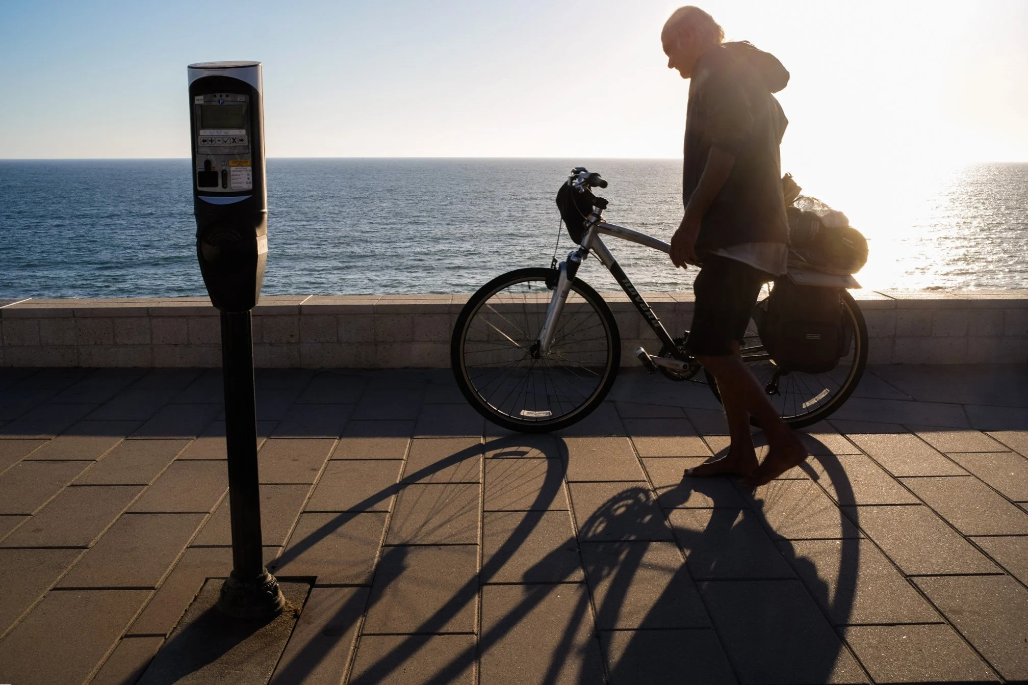 A man walking with a bicycle near a beach or ocean, with the sun setting or rising, casting shadows on the sidewalk and a parking meter in the foreground.