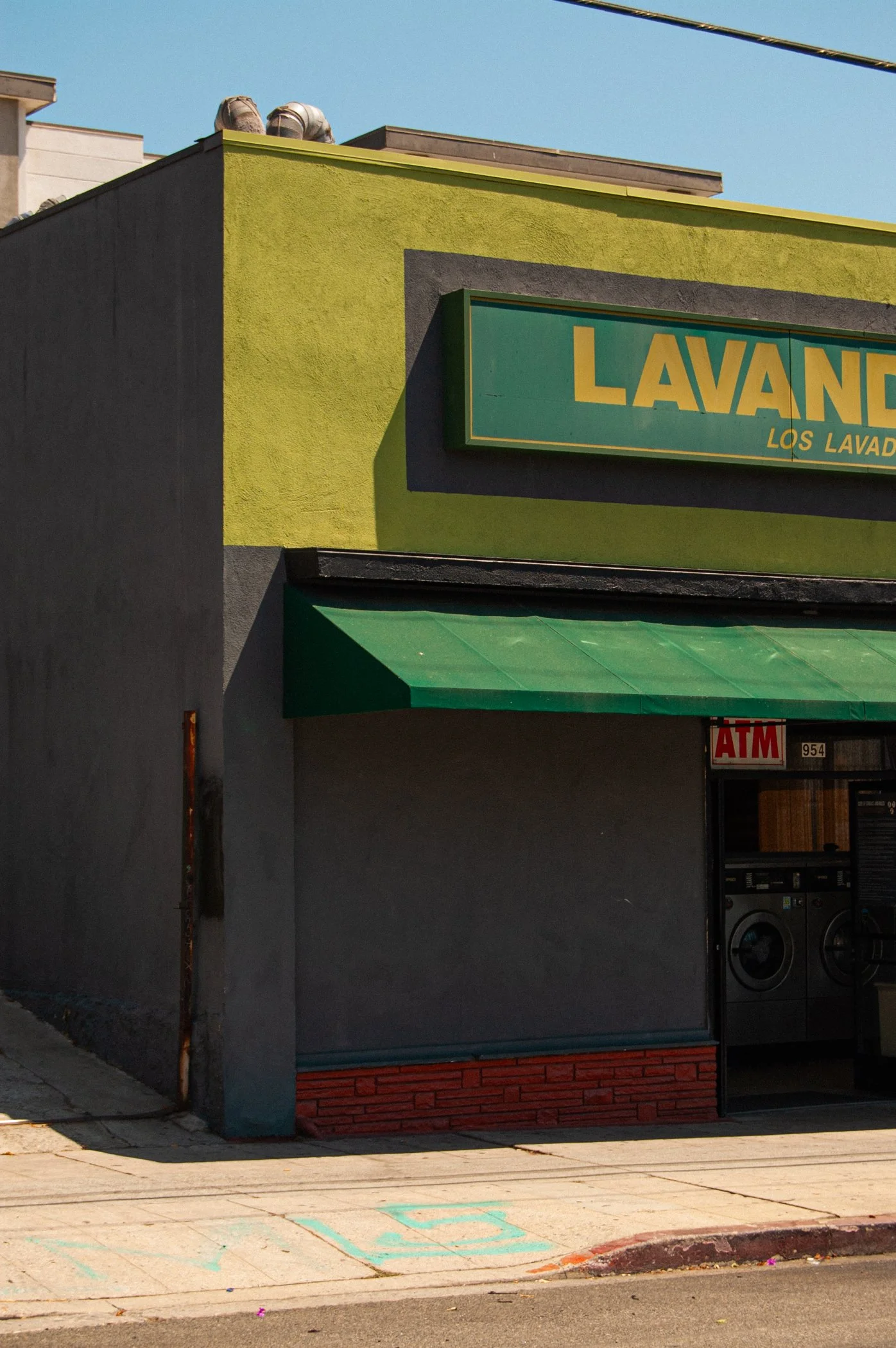 The image shows the exterior of a laundromat with a yellow and green sign that reads 'LAVAND' and 'LOS LAVAD', with laundry machines visible inside and a sidewalk in front of the store.