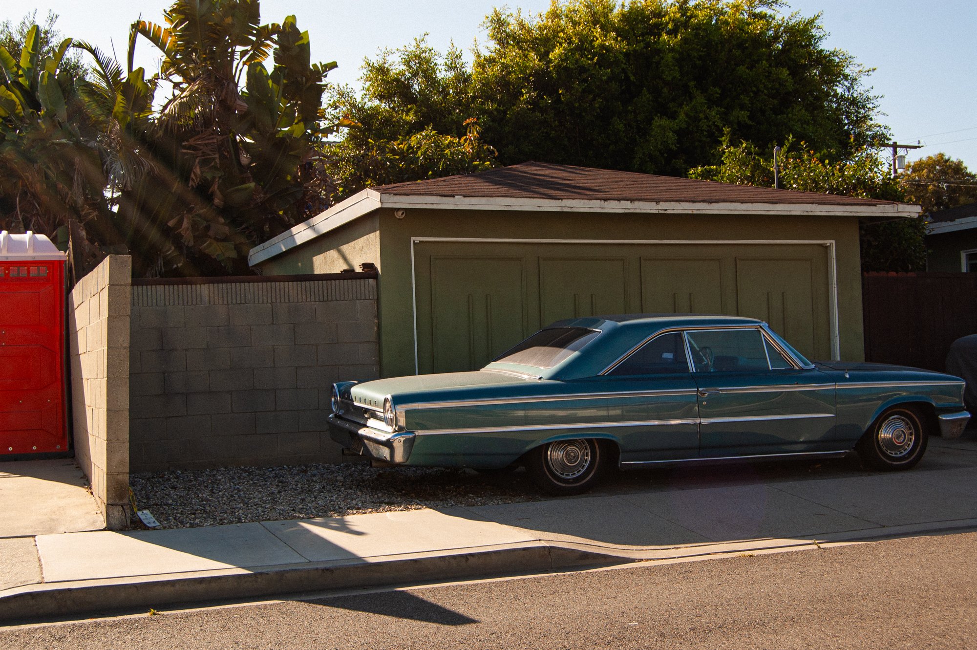 A vintage teal-colored car parked in front of a green garage with a sloped roof, next to a concrete block fence. A red mailbox is visible on the left side of the image, and large leafy trees are in the background. The scene is illuminated by sunlight