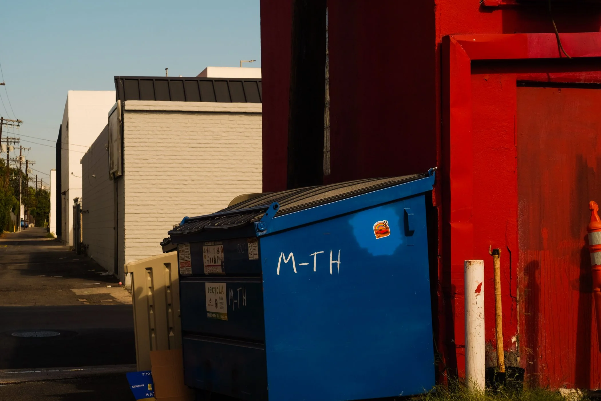 A row of colorful dumpsters in front of a red wall on a city street. The blue dumpster labeled "M-T-H" is prominently visible in the foreground, with a yellow dumpster behind it. In the background, there is a white building, utility poles, and wires.