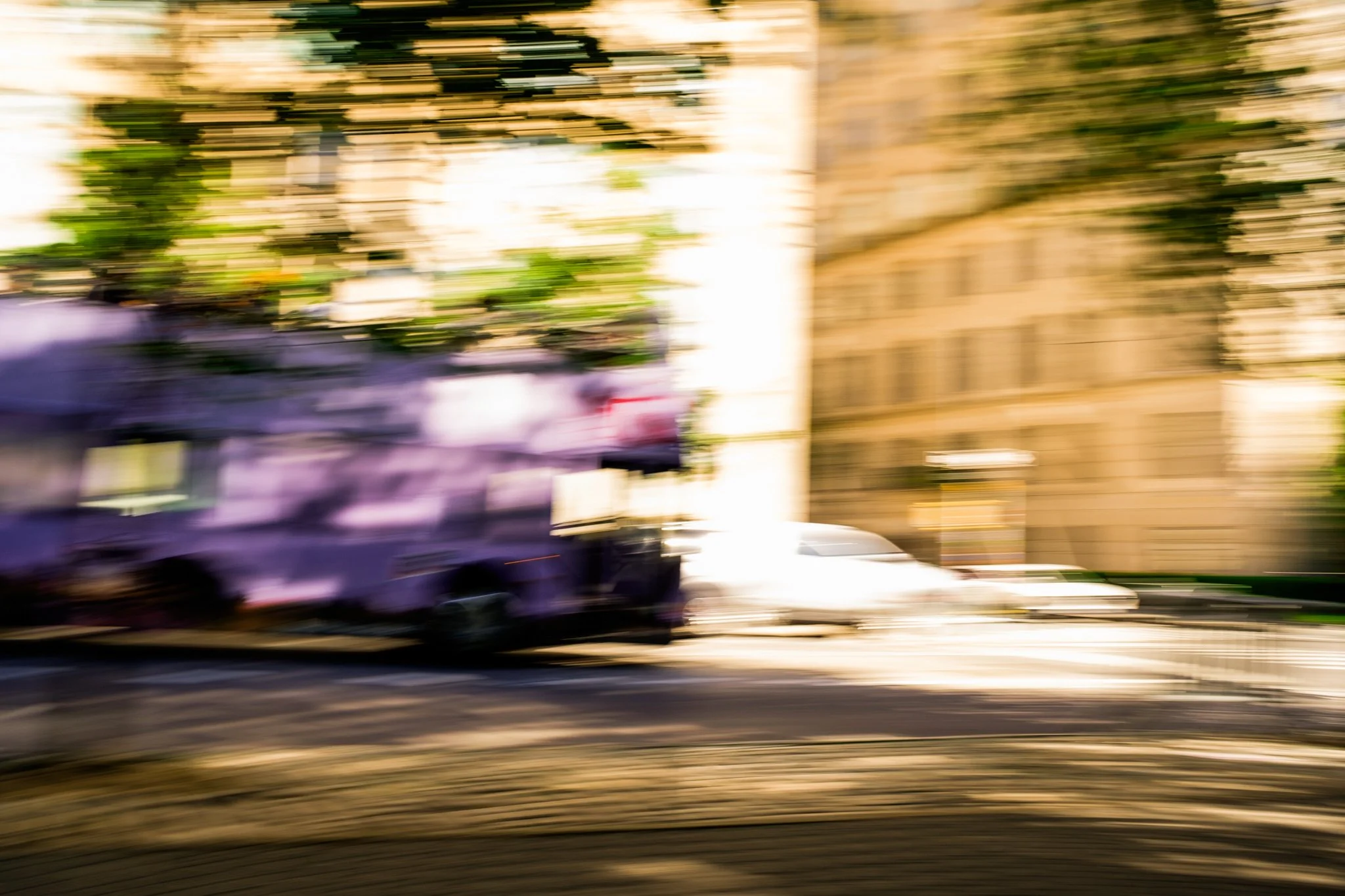 Blurred image of a vehicle and buildings, motion-filled scene with streaked trees and structures, sunlight in the background.