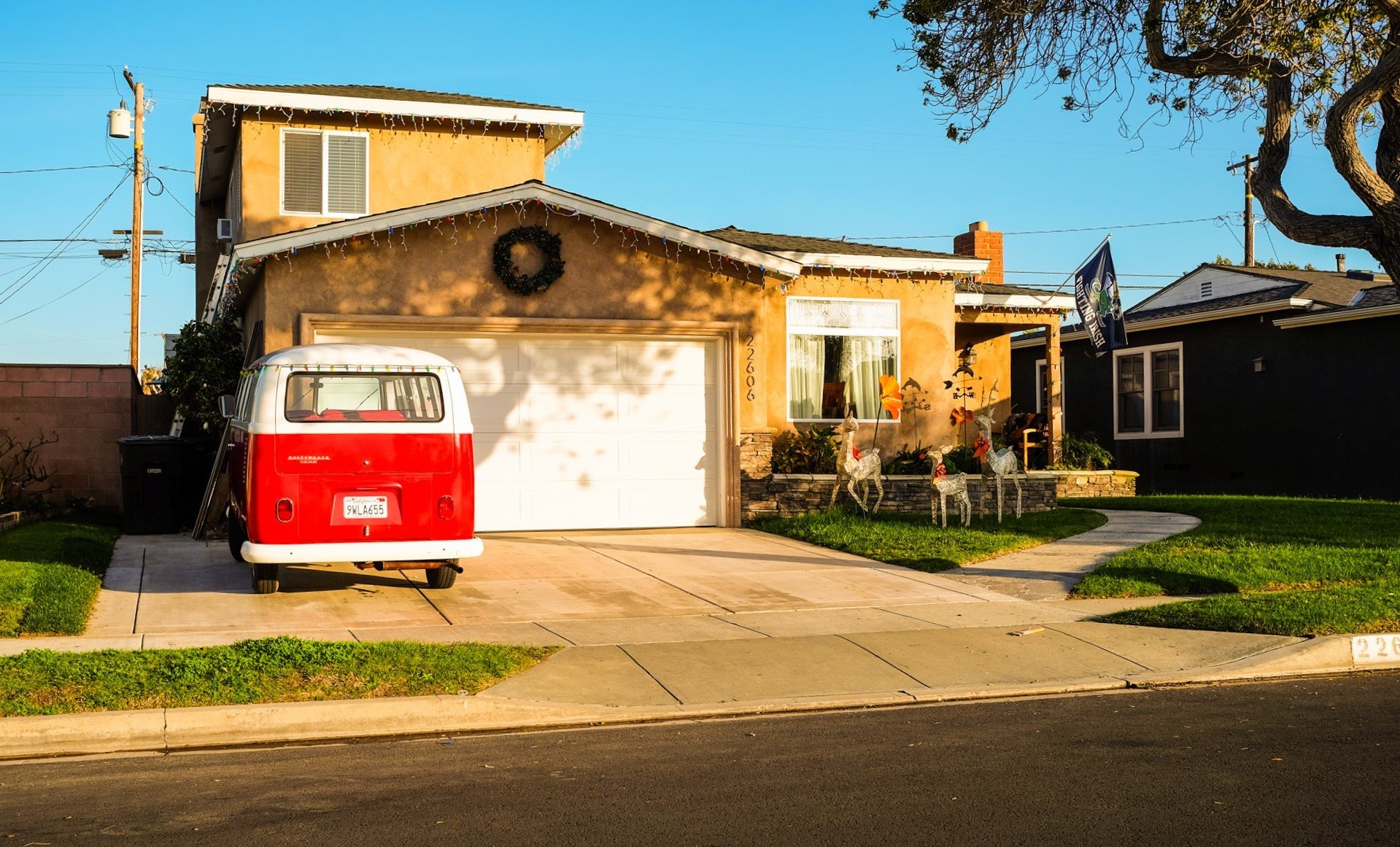 A two-story yellow house decorated with Christmas lights, with a red and white vintage van parked in the driveway, and holiday decorations including a wreath and reindeer figures in the front yard.