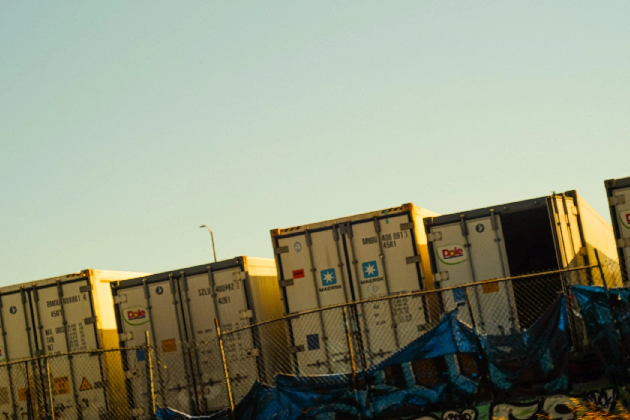 Stacked shipping containers in a lot under a light green sky, with a fence in the foreground and a streetlight in the background.