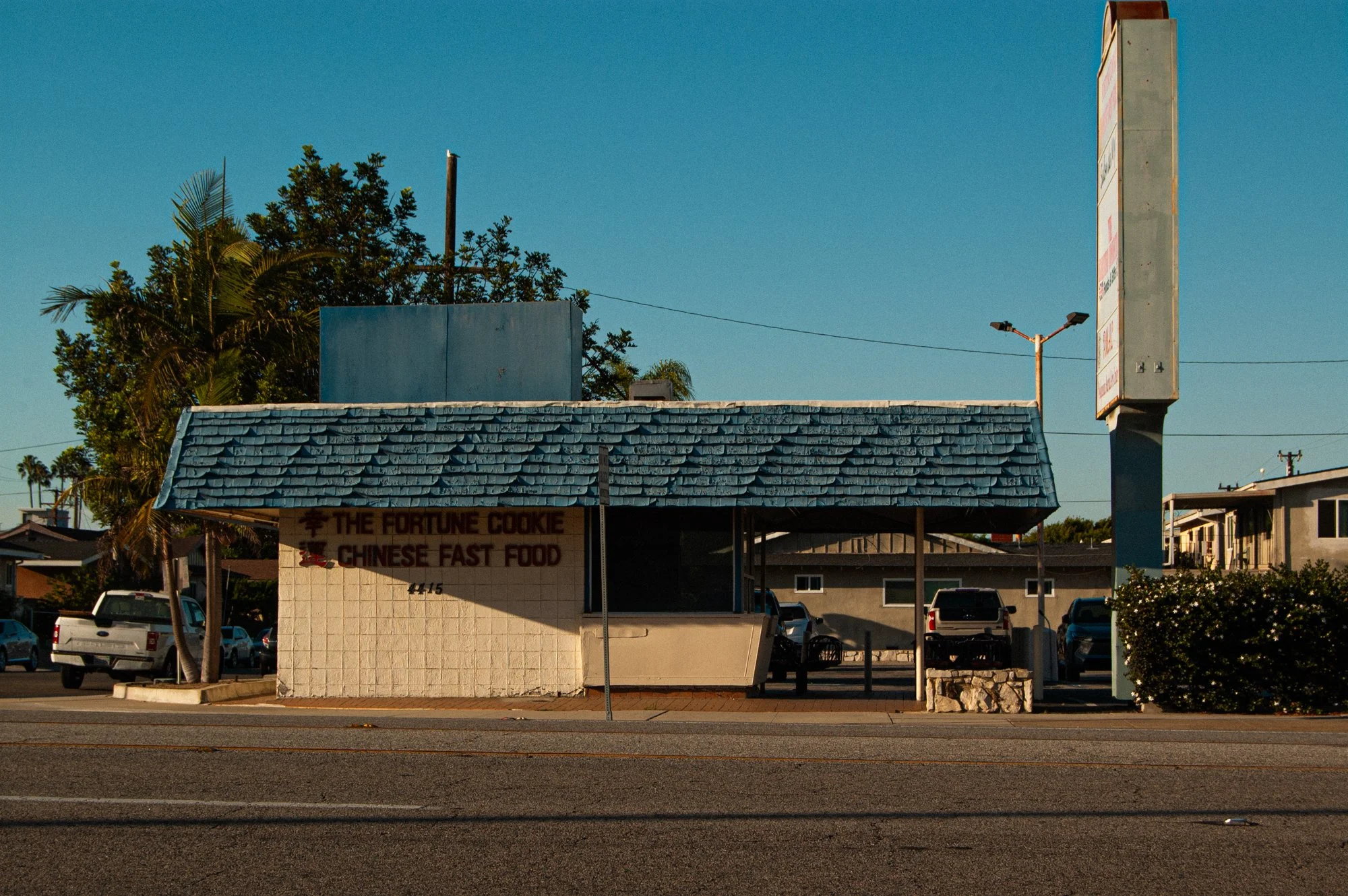 A small building with a sign that reads 'The Fortune Cookie Chinese Fast Food' on a sunny day. There are trees, parked cars, and a tall sign or billboard in the background.