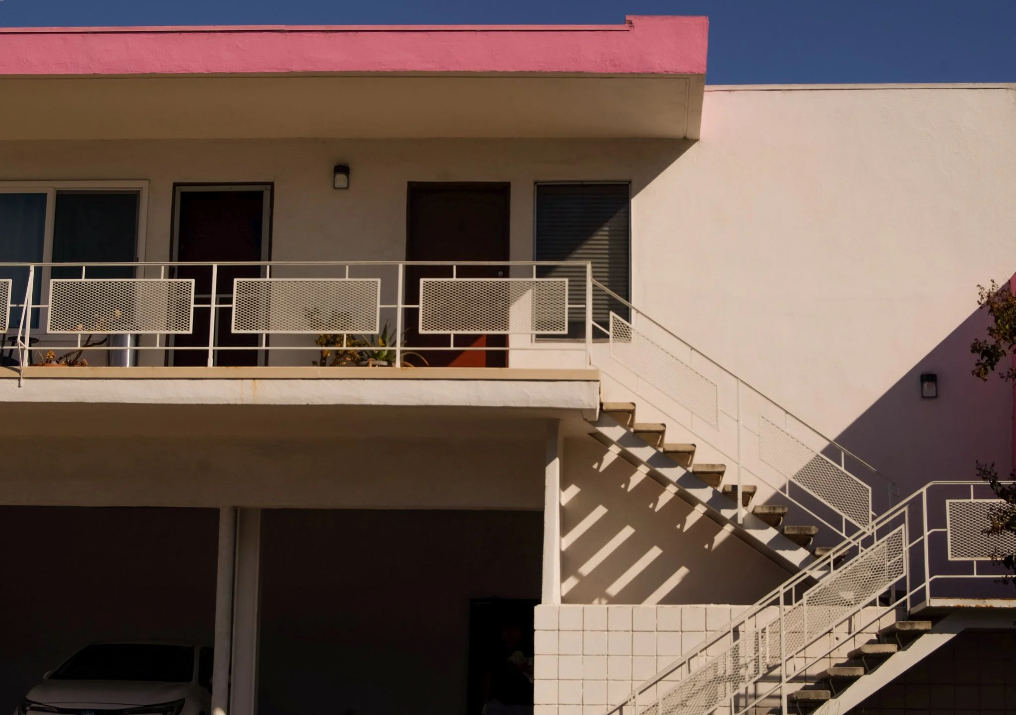 A multi-story building with a white exterior, pink upper section, and a white metal railing on the balcony. A staircase runs along the right side, casting diagonal shadows. A white vehicle is partially visible under the building, and the sky above is