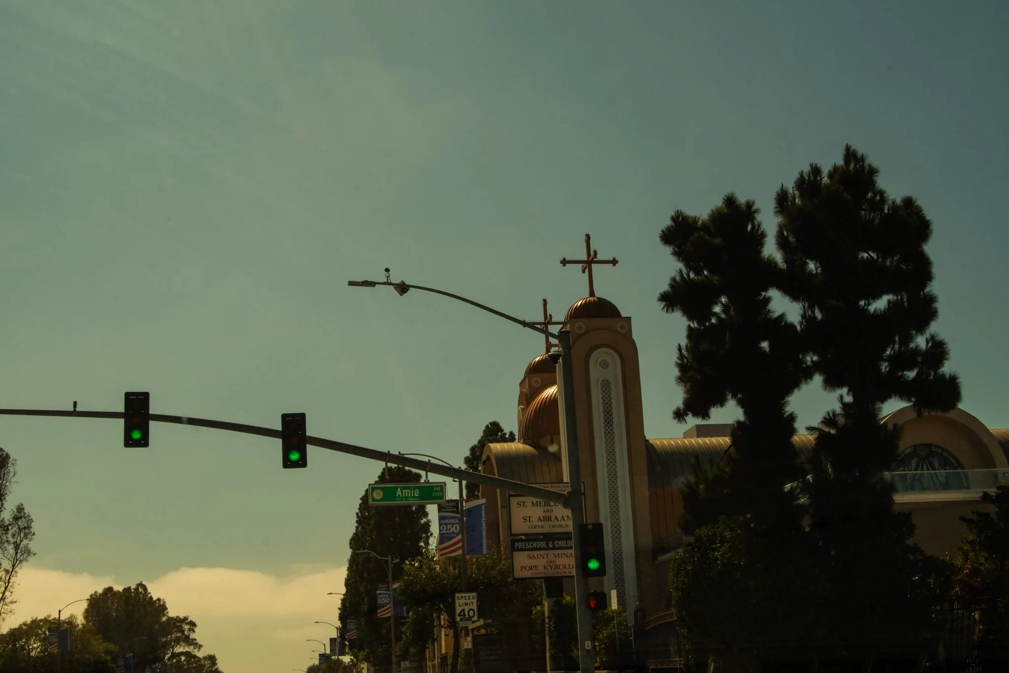 Street view showing traffic lights, street signs, church, and a large tree against a partly cloudy sky.
