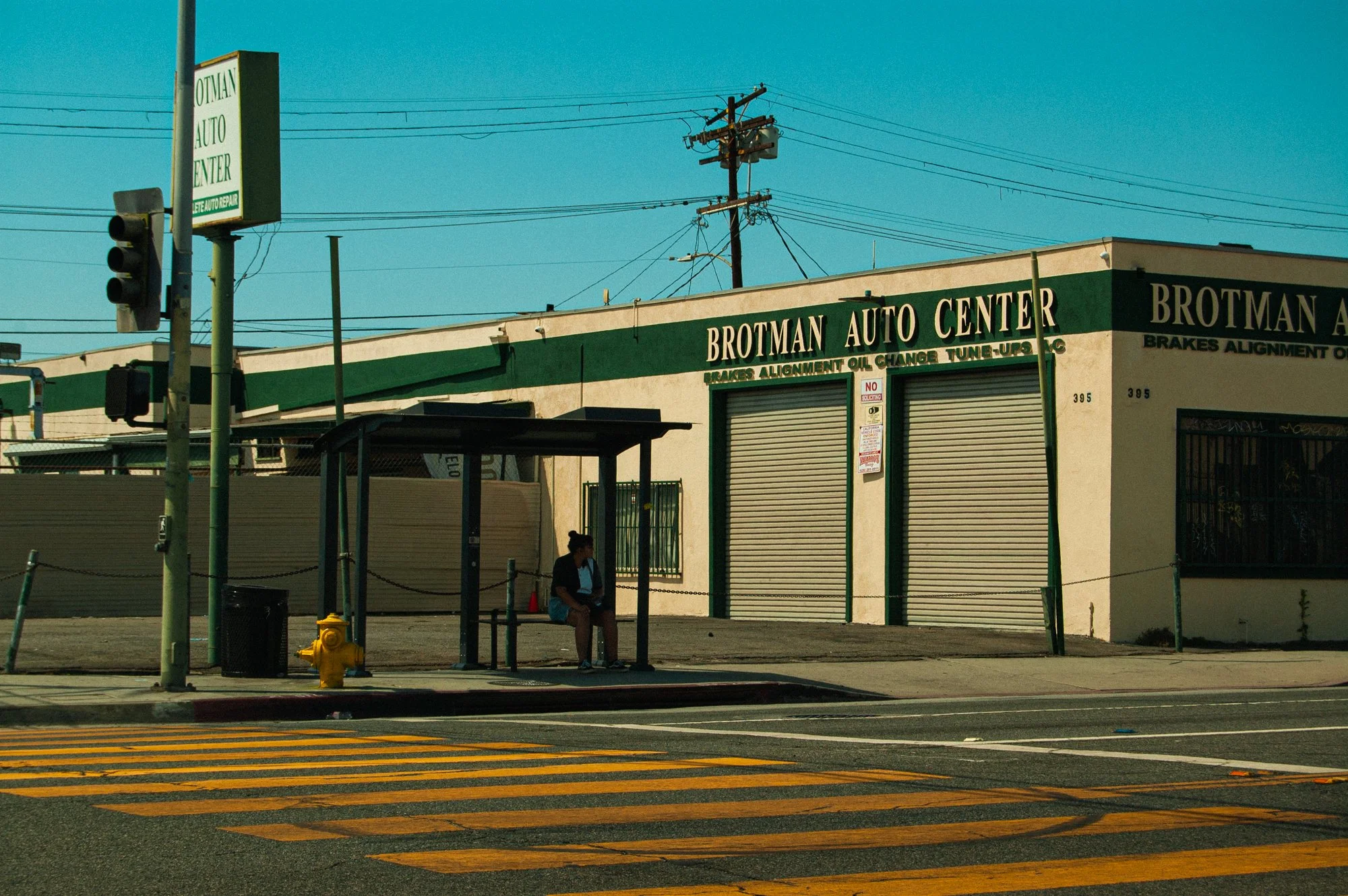 A woman sitting on a bench at a bus stop in front of a green auto repair shop called Brotmans Auto Center, with a bus shelter, a bus stop sign, and a traffic light.