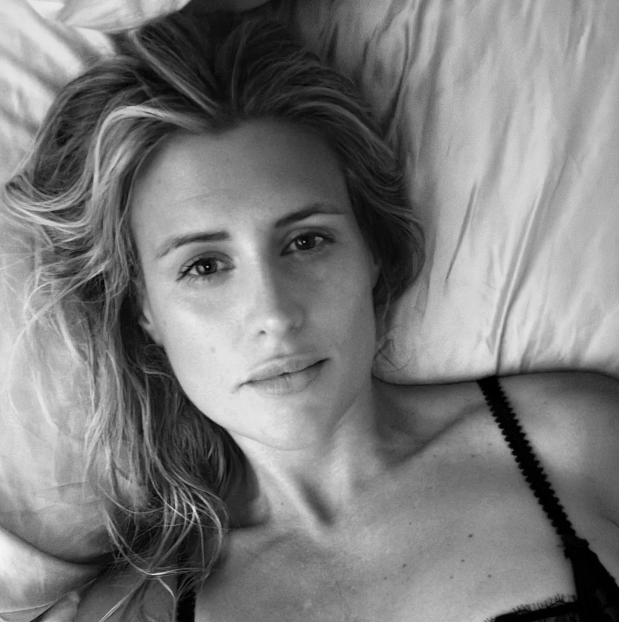 A black and white photograph of a young woman, Candace Max, wife of musician Kevin Max, lying down, looking directly at the camera with a relaxed expression, with tousled hair and a black strap visible on her shoulder.