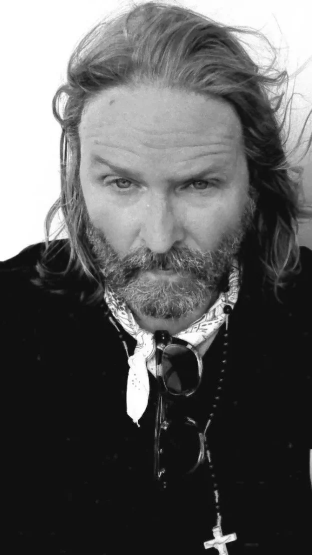Black and white close-up photo of artist and Grammy award winning musician Kevin Max with long, wavy hair and a beard, wearing sunglasses hanging from a patterned bandana or scarf around his neck, and a cross necklace.