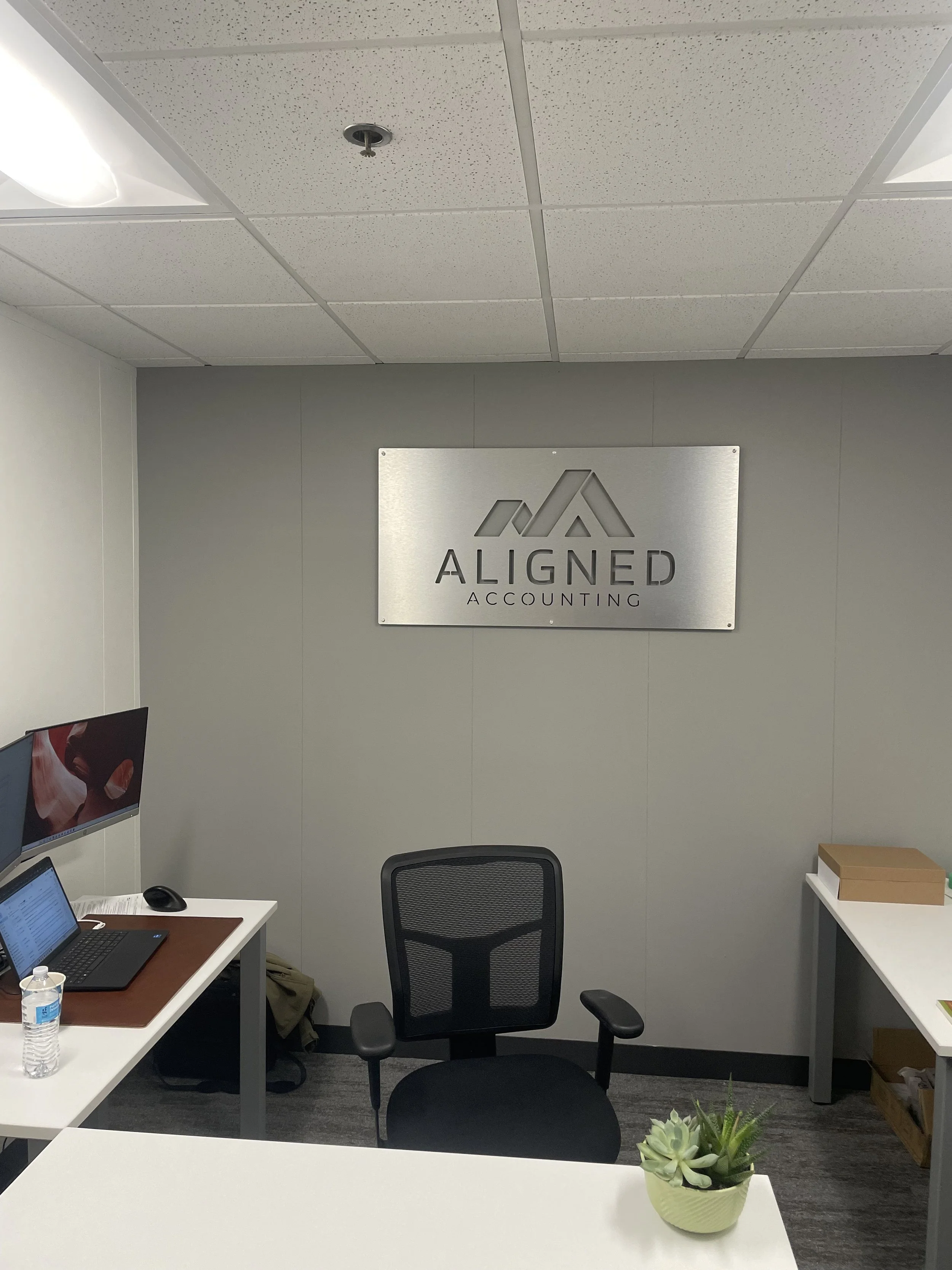 Office space with a desk, computer, water bottle, and a succulent plant in a room with gray walls and a sign that reads 'Aligned Accounting' on the wall.