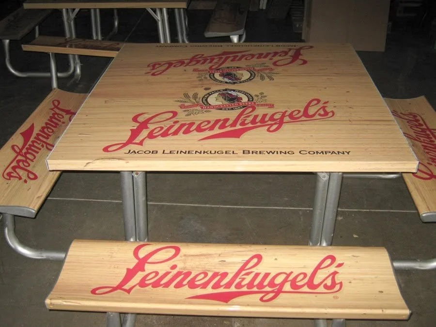 Wooden table and benches with Leinenkugel's beer branding, located in a brewery or pub setting.