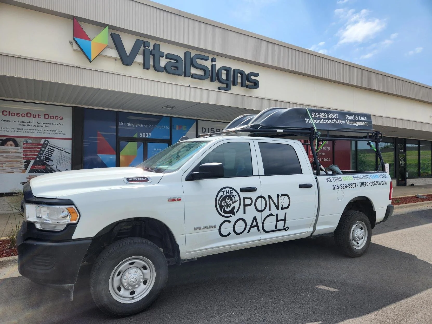 White RAM pickup truck with 'The Pond Coach' logo and text parked outside a Vital Signs store. The truck has a rack on the back with signs advertising pond and lake management services, including website and phone number. The store has large windows 