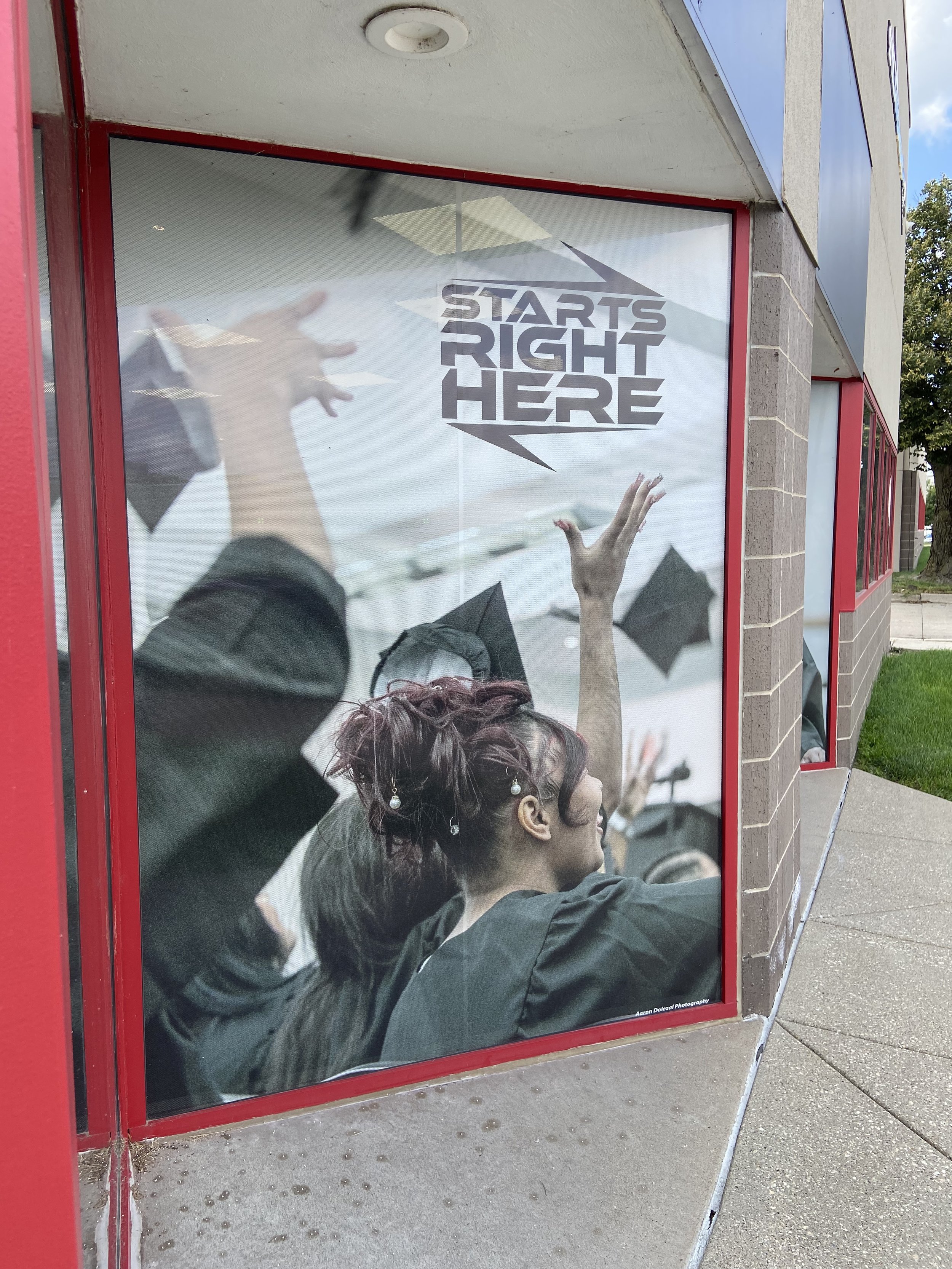 Reflective glass window with the phrase 'Starts Right Here' and a large hand reaching upward, surrounded by other hands in various positions, with a brick building corner and sidewalk outside.