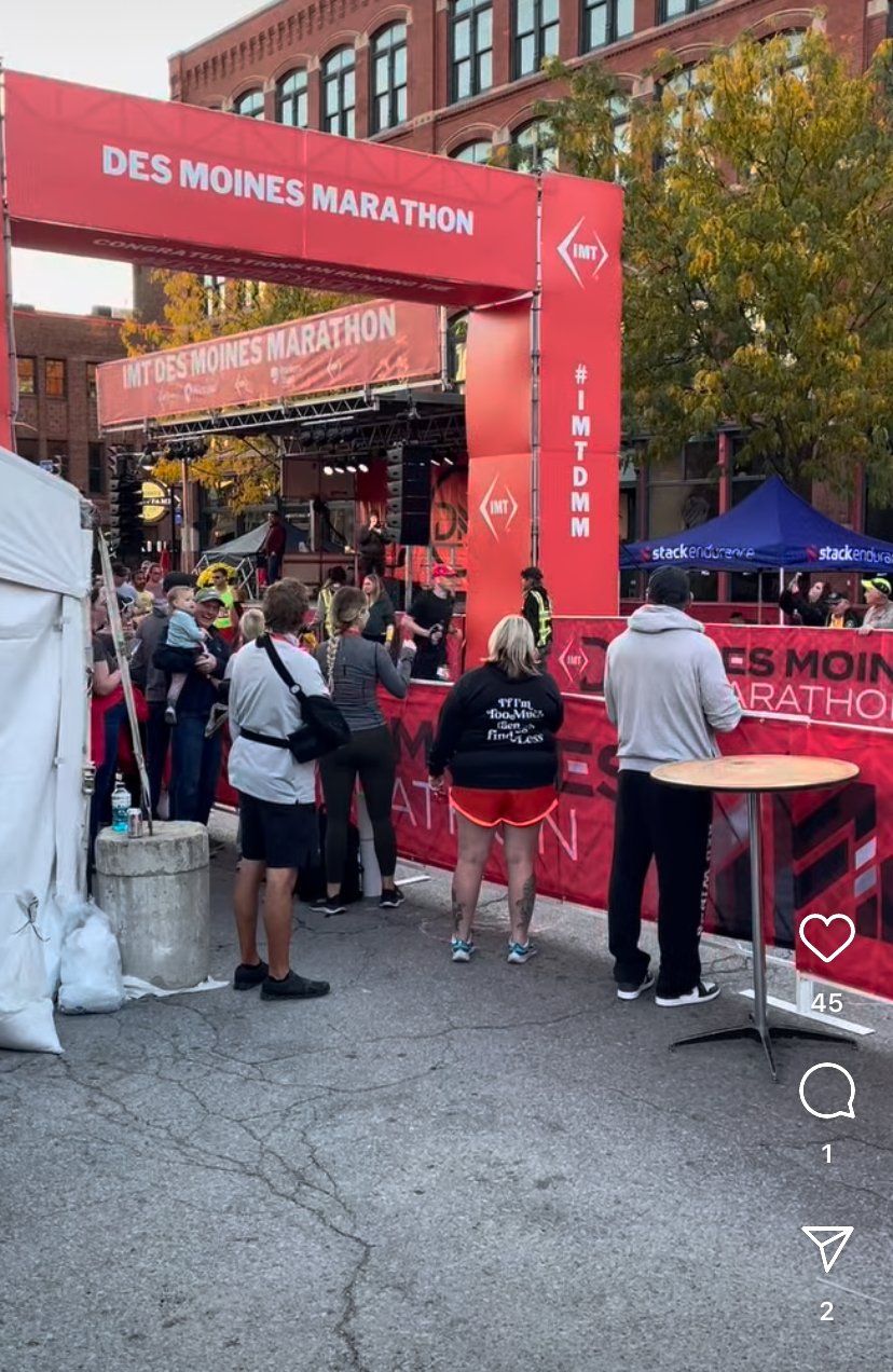 People gathered in front of a stage at the Des Moines Marathon in Iowa, with a red banner overhead, surrounded by tents and trees with autumn foliage.