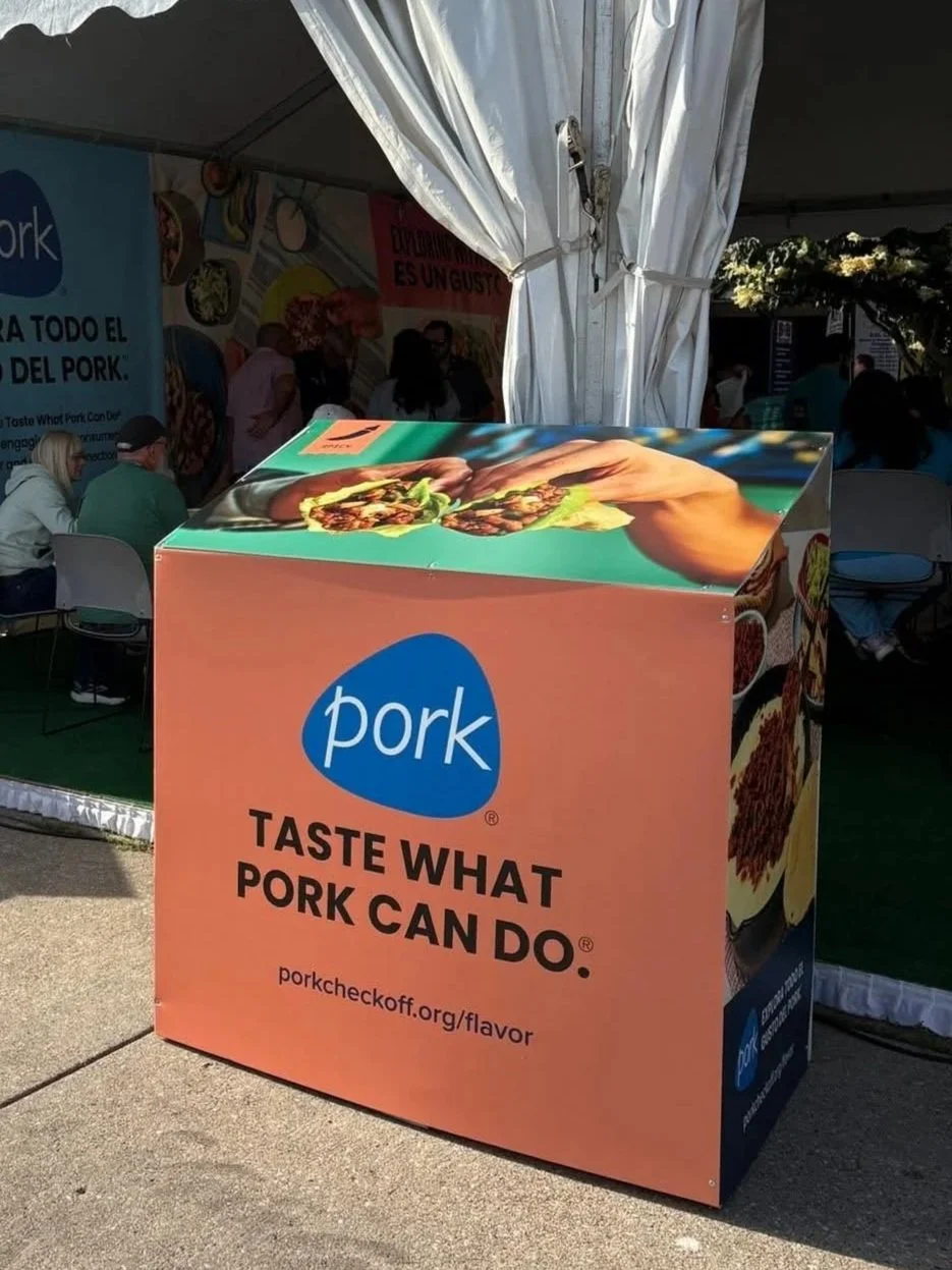 Outdoor booth promoting pork with a sign reading "Taste what pork can do" and a website link, surrounded by people and banners.