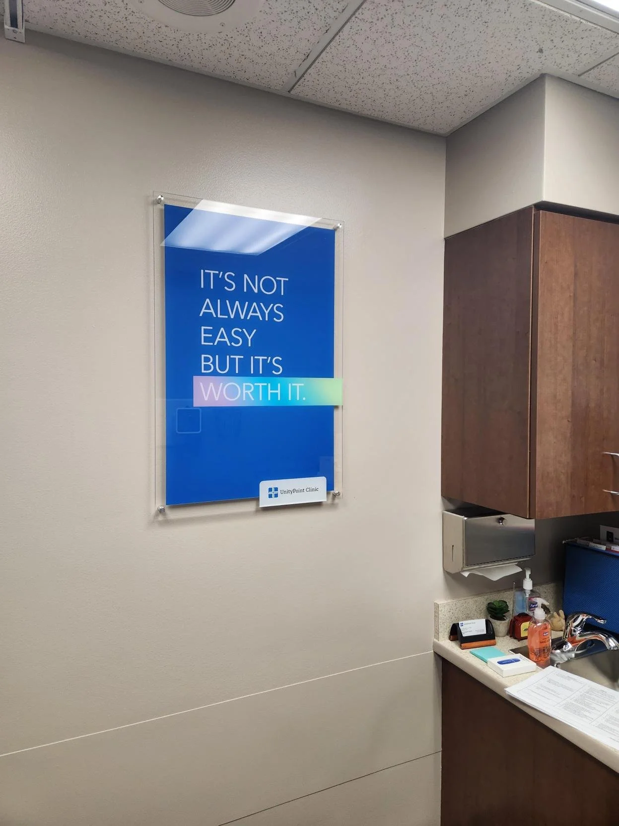 Interior view of a doctor's office or clinic room with a motivational acrylic print on the wall. The acrylic print reads: "It's not always easy but it's worth it." There is a counter with a sink, hand sanitizer, paper, and medical supplies. A wooden 