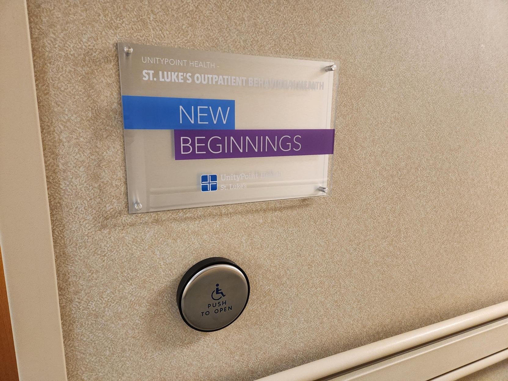 Acrylic wayfinding on beige wall reading "UnityPoint Health - St. Luke's Outpatient Behavior" with blue and purple boxes stating "New Beginnings." A smaller sign below indicates accessibility with a wheelchair symbol and says "Push to open."